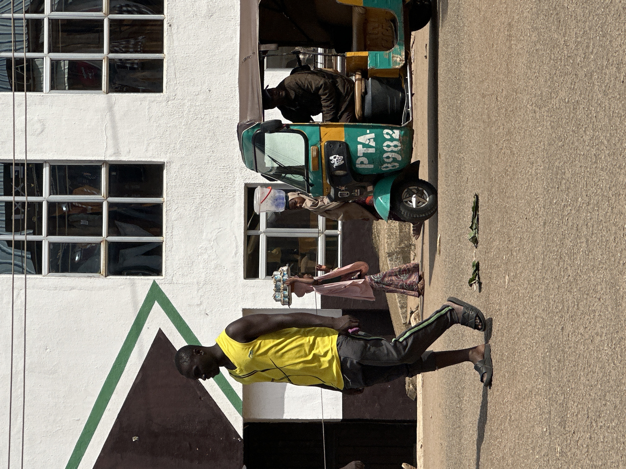 In Bukuru, Nigeria, a man in a bright yellow tank top and dark pants walks across an asphalt street. He wears dark slides and carries a purple object in his right hand. Behind him, a woman wearing a headscarf and a patterned dress balances a tray of items on her head. To her right, a turquoise and yellow auto-rickshaw, marked "PTA-8982," is parked. A man in a dark jacket sits in the driver's seat of the auto-rickshaw. A woman in a light-colored headscarf stands beside the auto-rickshaw with a red and white bucket on her head. The background features a white building with dark brown and green triangular accents, and multiple windows.