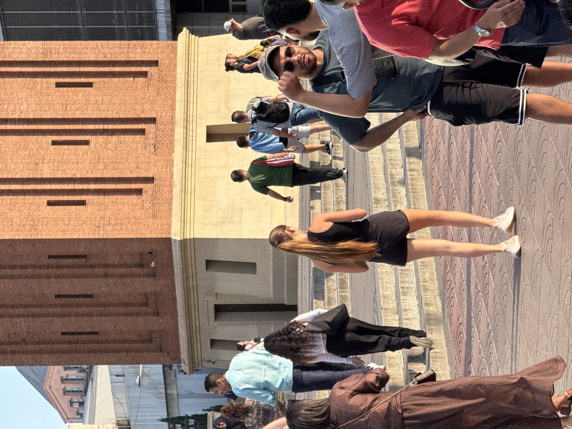 This image captures a street scene in Barcelona, Spain, near the Arenas de Barcelona, a former bullring that has been converted into a shopping center and entertainment venue. In the foreground, several people are walking and standing. On the left, a woman in a brown dress walks away from the camera. To her right, a woman with long, dark curly hair in a white crop top and black pants carries a black shoulder bag. In the center, a woman with a blonde ponytail wears a black tank top and black shorts.
Further back, a man in a light blue shirt and dark pants walks towards the left. A group of men are also visible, including one in a green t-shirt, another in a blue t-shirt and shorts, and one with a backpack wearing a striped shirt. In the mid-ground, two women talk, and a man in a gray t-shirt and sunglasses stands with his hand raised, talking to another man wearing a red shirt.
The background features a large, red brick tower with distinctive vertical indentations, characteristic of the architecture surrounding the Arenas. The ground is paved with a patterned brick walkway. Sunlight casts shadows, indicating it is likely late afternoon or early evening.
