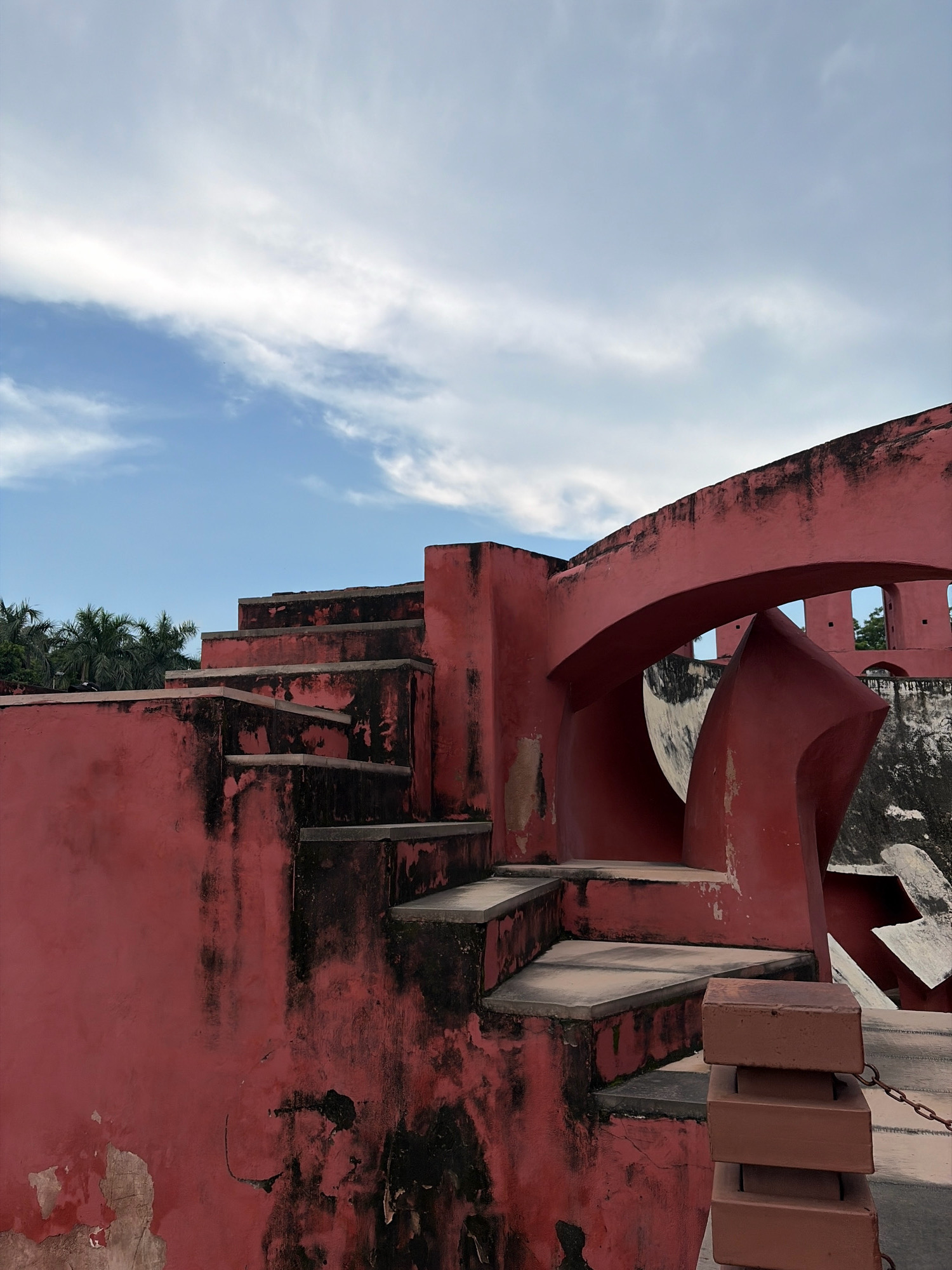The image shows the Samrat Yantra, a monumental astronomical instrument located at the Jantar Mantar in New Delhi, India. The structure is made of red sandstone and features a large gnomon that casts a shadow to tell time. The weathered pink walls of the Samrat Yantra are visible, with dark patches indicating age and exposure. A set of concrete stairs ascends along the side of the structure. In the background, lush green palm trees are visible against a bright blue sky with scattered white clouds. The scene is devoid of people.