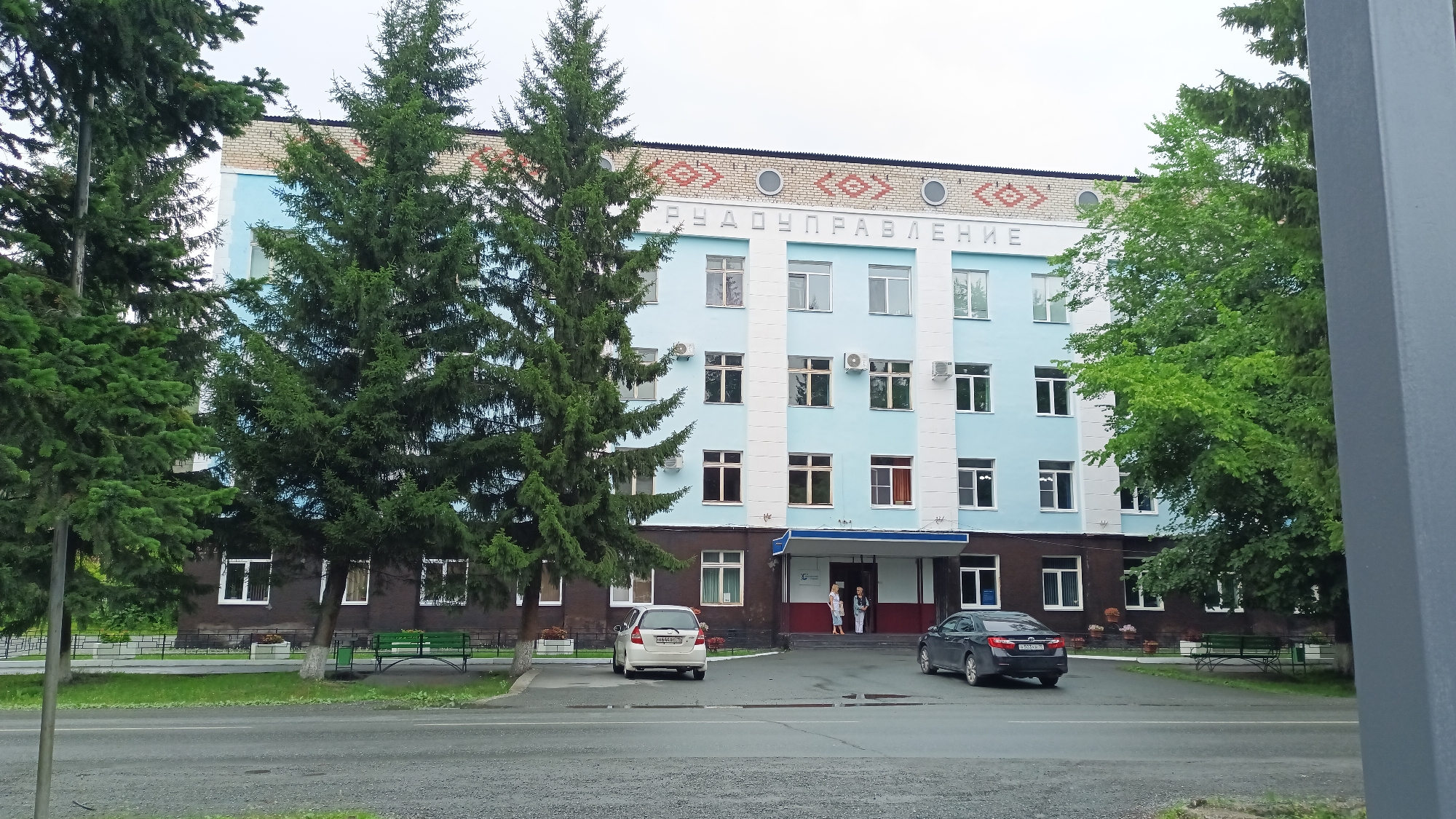 This media file shows a light blue, multi-story building with white vertical accents. Above the windows on the top floor, there is decorative brickwork with a patterned border and circular elements. Cyrillic text is visible on the building, spelling out "РУДОУПРАВЛЕНИЕ", which translates to " Ore Administration" or "Mine Administration". The building appears to be in a public area, possibly a town or city. In front of the building, there is a paved area with two cars parked. One is a light-colored hatchback, and the other is a dark sedan. Two people are standing near the entrance of the building.

Flanking the building are several tall evergreen trees and deciduous trees with lush green foliage, suggesting a temperate climate. In the foreground, there is a road with a grey asphalt surface. The sky is overcast, indicating a cloudy or possibly rainy day. Green benches are visible along a walkway in front of the building, and some planters with flowers are also present. The overall scene suggests a daytime setting in Abaza, Russia, likely a functional administrative building surrounded by a green, public space. The mood is neutral, with no strong emotional cues.
