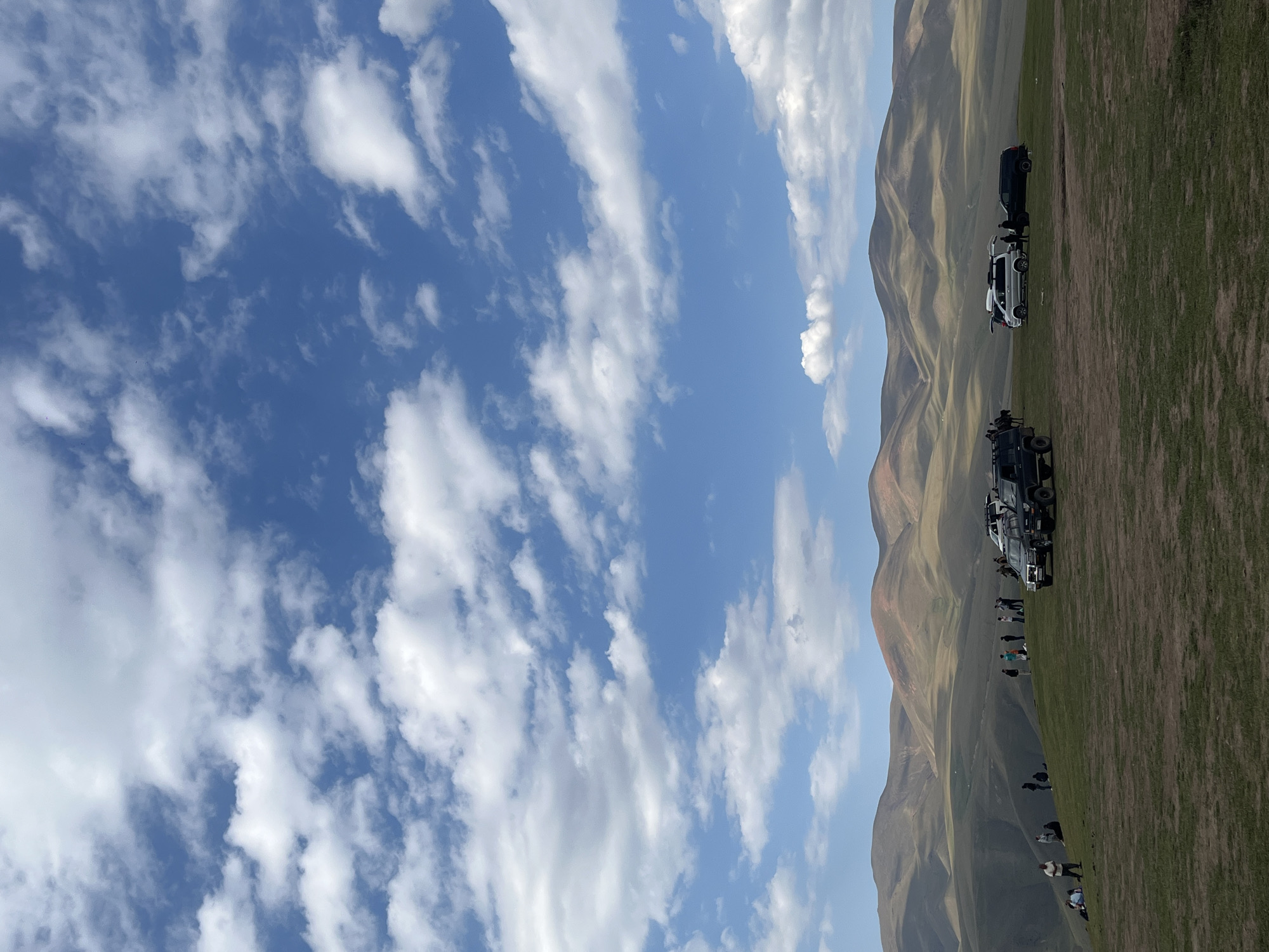 The image depicts a wide shot of a grassy, rolling landscape under a bright blue sky with scattered white clouds. In the foreground, a group of people are gathered on the hillside, some standing, some sitting. Several vehicles are parked on the grass, including two dark-colored SUVs, a white minivan, and another dark SUV. In the middle ground, more people are visible in small groups, and a horse is also present. The background features a series of undulating hills and mountains, with some areas showing reddish-brown patches. The scene appears to be a popular gathering spot in the outskirts of Batan, Kazakhstan, possibly for recreation or sightseeing, given the presence of vehicles and the scenic vista.