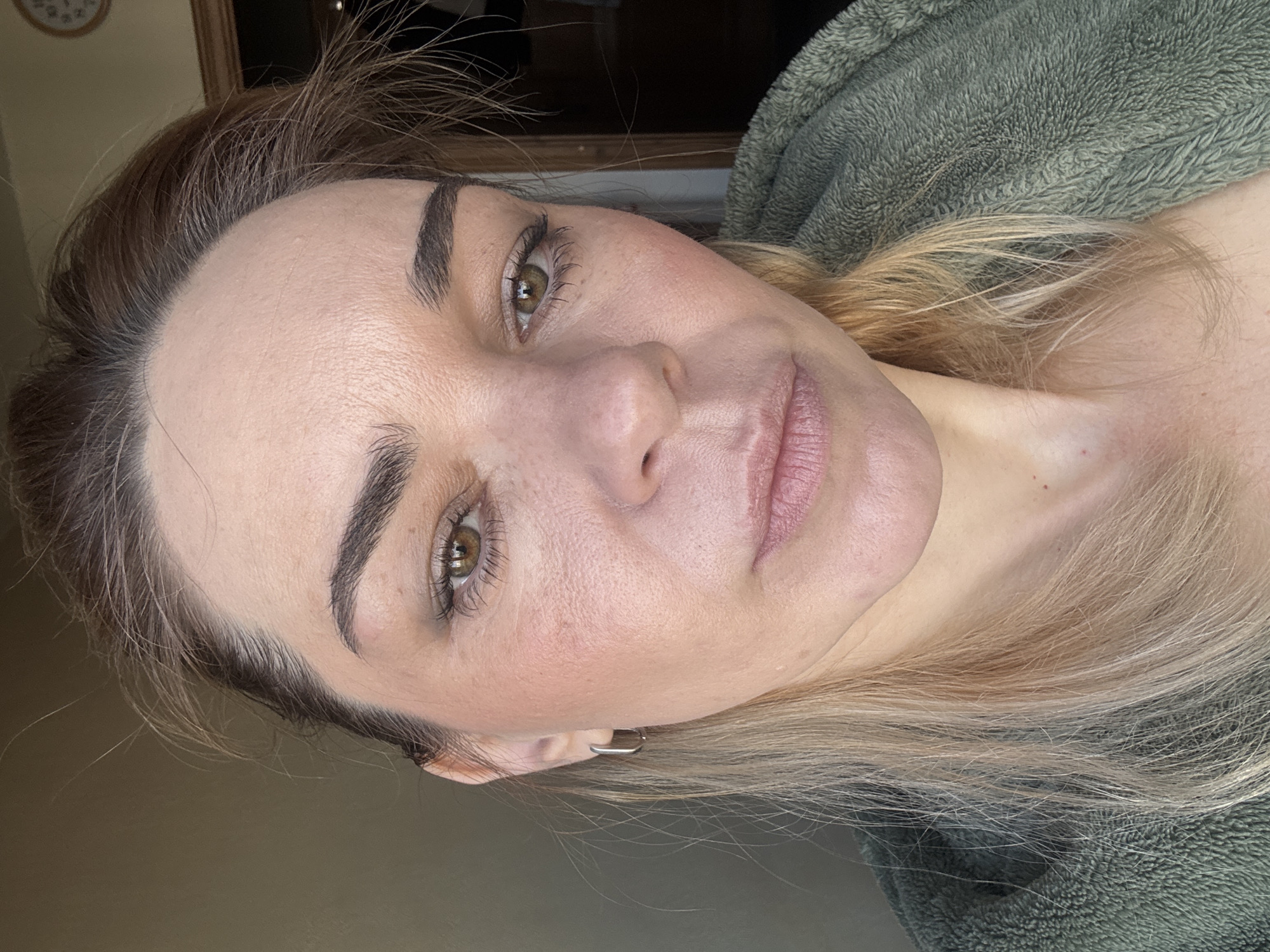 A close-up, slightly tilted portrait depicts a woman with fair skin and light blonde and brown hair. She has hazel eyes and well-defined eyebrows. Her face appears to have minimal makeup, with a light blush on her cheeks and a subtle lip color. She is wearing a small silver hoop earring. The background is blurred, showing a portion of a wall and a dark green, possibly soft fabric. No specific details indicate the location as Unknown, Russia.