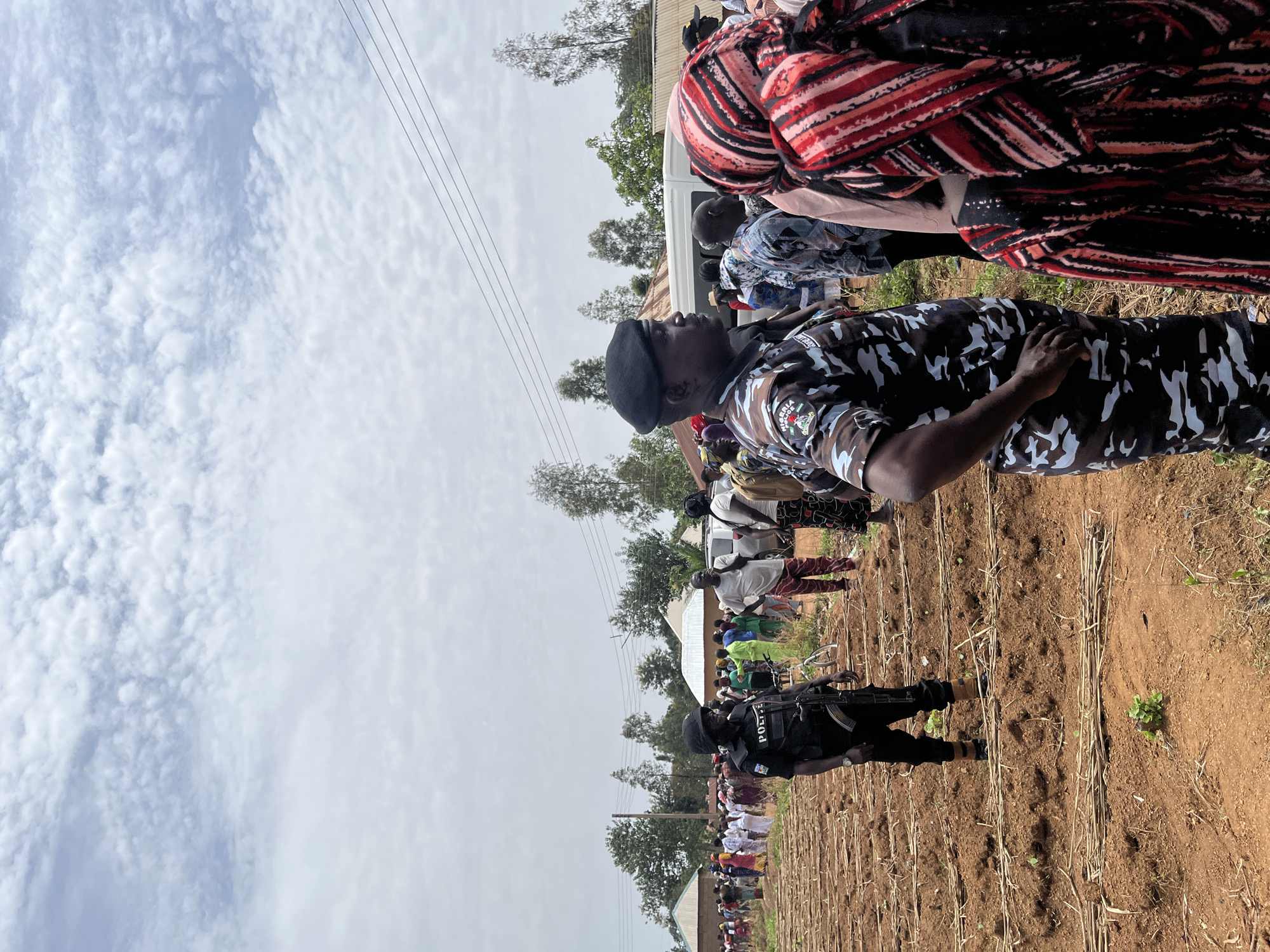 In Dutsen Kato, Nigeria, a large group of people stands in a field of dry stalks with a police presence.
People: The crowd consists of men, women, and children, some wearing colorful clothing, others in more subdued attire. Two police officers are visible: one in camouflage fatigues with a beret stands near the front of the crowd, while another in full black gear with "POLICE" emblazoned on their vest stands guard further back.
Environment: The scene takes place in a field of dry stalks in a dirt field, suggestive of recently harvested crops. A few trees are visible in the background, along with a building. The sky is filled with clouds. Overhead power lines extend into the distance.
Actions: The individuals in the crowd are mainly standing or milling about. The police officers appear to be keeping watch over the crowd.