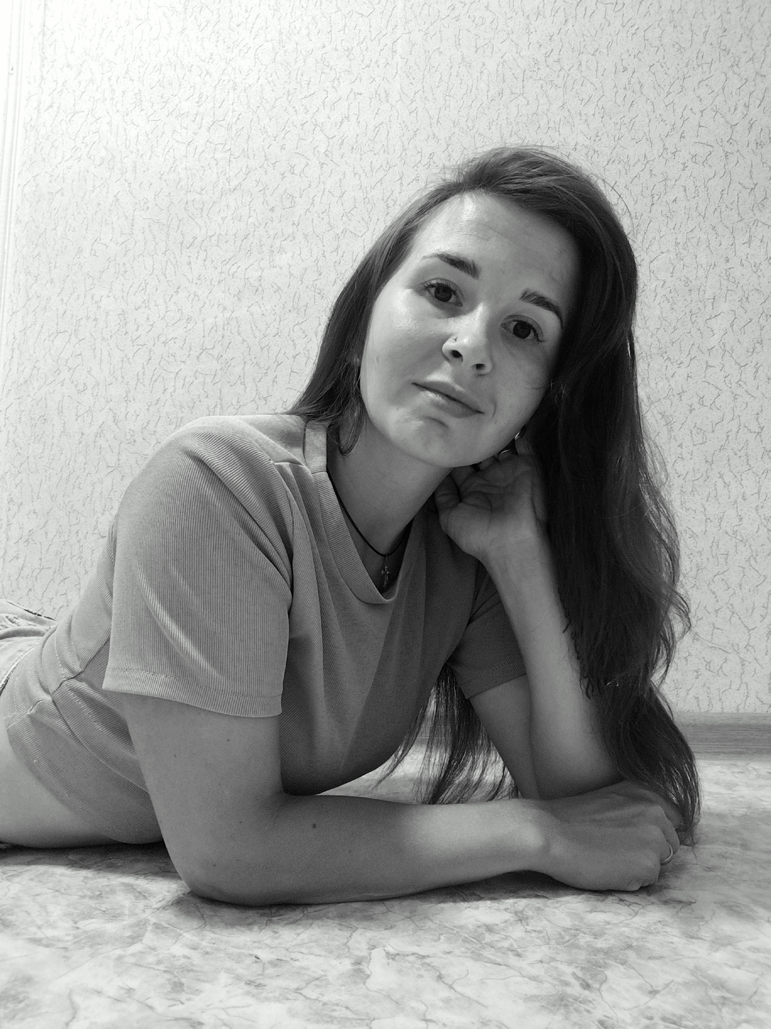 A black and white, medium shot photo shows a young woman lying on the floor. She has long, dark hair that falls around her shoulders and down her back. Her head is propped up on her hand, and she is looking directly at the camera with a relaxed expression. She is wearing a t-shirt and shorts. The background is a textured wall. The floor appears to be tiled with a marble-like pattern. The lighting suggests it is daytime, possibly indoors. There is no visible text in the image. The woman is the sole subject of the photograph.