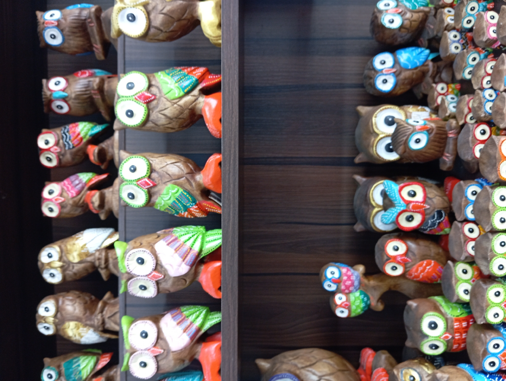 The image displays a shelf filled with a collection of carved wooden owls, likely a souvenir display in a shop in Nha Trang, Vietnam. The owls vary in size, color, and design, with many featuring bright, patterned wings and large, circular eyes. They are arranged on two shelves against a dark, wooden background. The upper shelf contains larger owls, some with visible feet painted orange or yellow. The lower shelf holds smaller owls, some clustered together, and a few larger ones with intricate detailing. The lighting appears to be from above, casting some shadows. The overall impression is one of a display of handcrafted decorative items.
