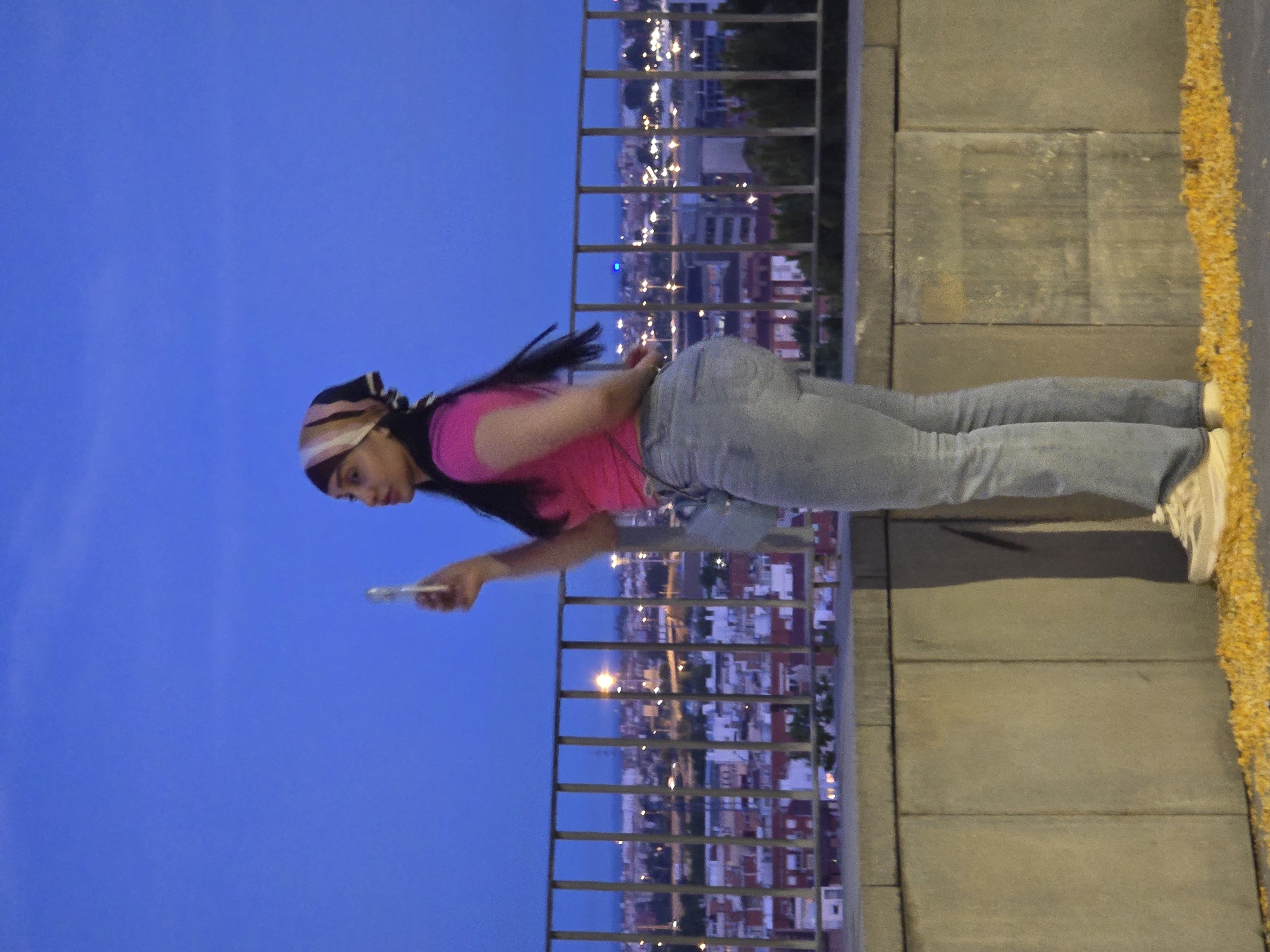 A woman stands on a stone wall overlooking a city at dusk. She is wearing a pink t-shirt, light blue jeans, and a patterned headscarf. She holds a mobile phone in her right hand, as if taking a picture or recording a video. In the background, the city lights of Cornellà de Llobregat, Spain, are visible against a darkening blue sky. A metal railing separates the woman from the cityscape. Yellowish foliage is scattered on the ground in the foreground.