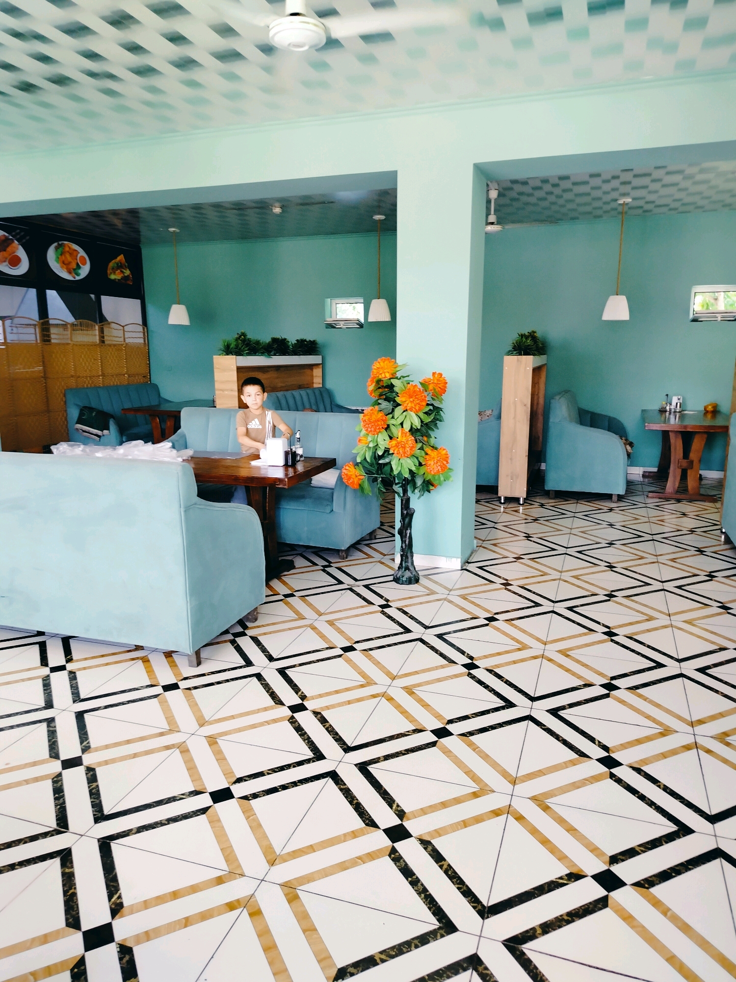 A boy is seated at a table in a modern cafe with teal walls and a geometric tiled floor. He is wearing a brown t-shirt and blue jeans and appears to be looking at something on the table. The cafe is furnished with light blue upholstered booth seating and dark wood tables. An artificial orange tree is positioned to the left of the frame. In the background, there are windows, ceiling fans, and pendant lights. A sign with images of food is visible on the left side of the image. The cafe is located in the city of Kölkent, Kazakhstan.