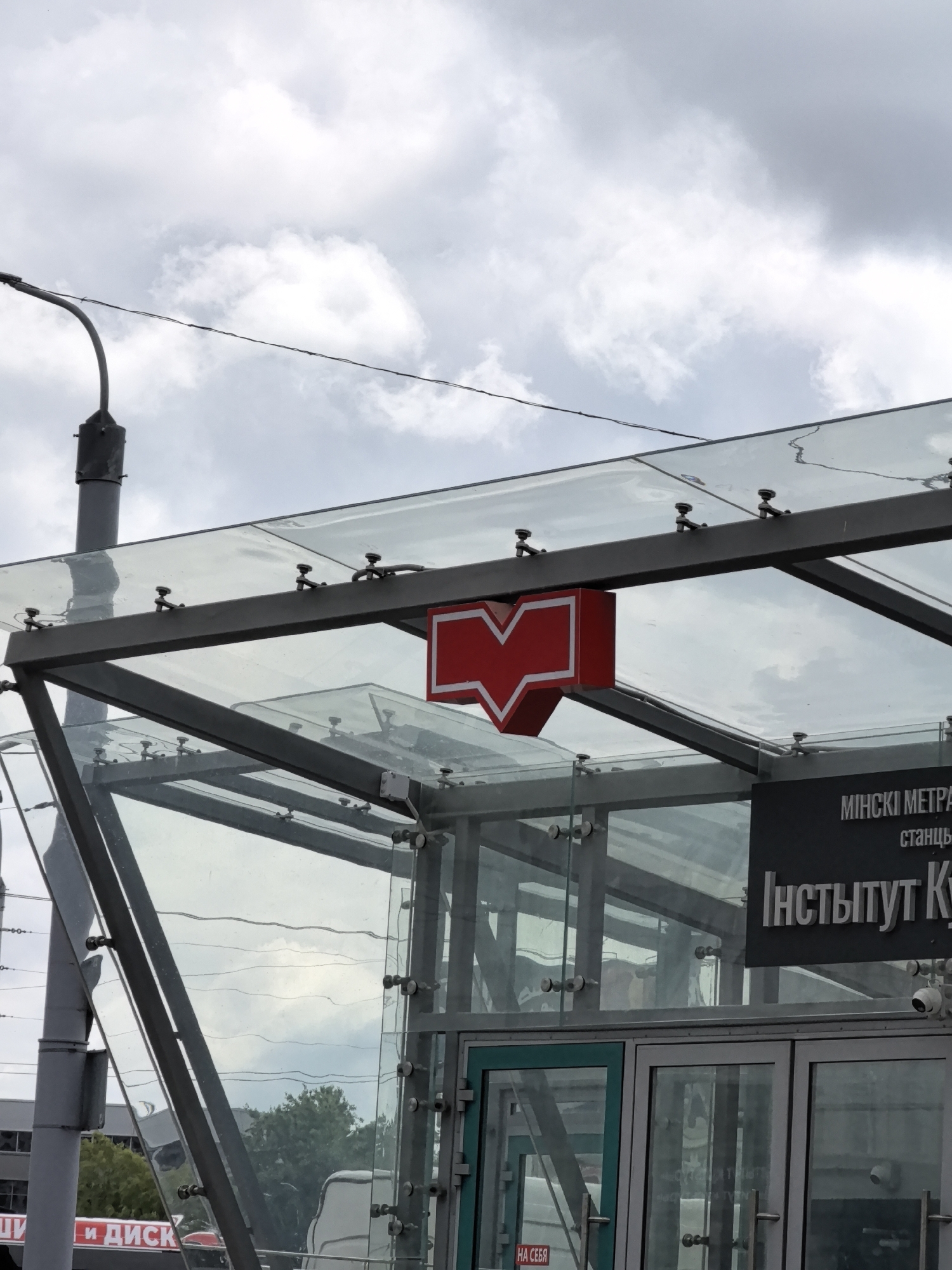 A red and white metro station logo is visible on a glass structure under a cloudy sky. A metal pole is to the left of the structure, and a sign with Cyrillic text is mounted on the right. The sign appears to say "МІНСКІ МЕТРА" and "Інстытут К". Part of a red and white advertisement with Cyrillic text, possibly reading "ЦИ И ДИСК", is visible at the bottom left. The structure features a glass roof supported by metal beams and glass walls. Security cameras are also visible on the exterior of the structure.