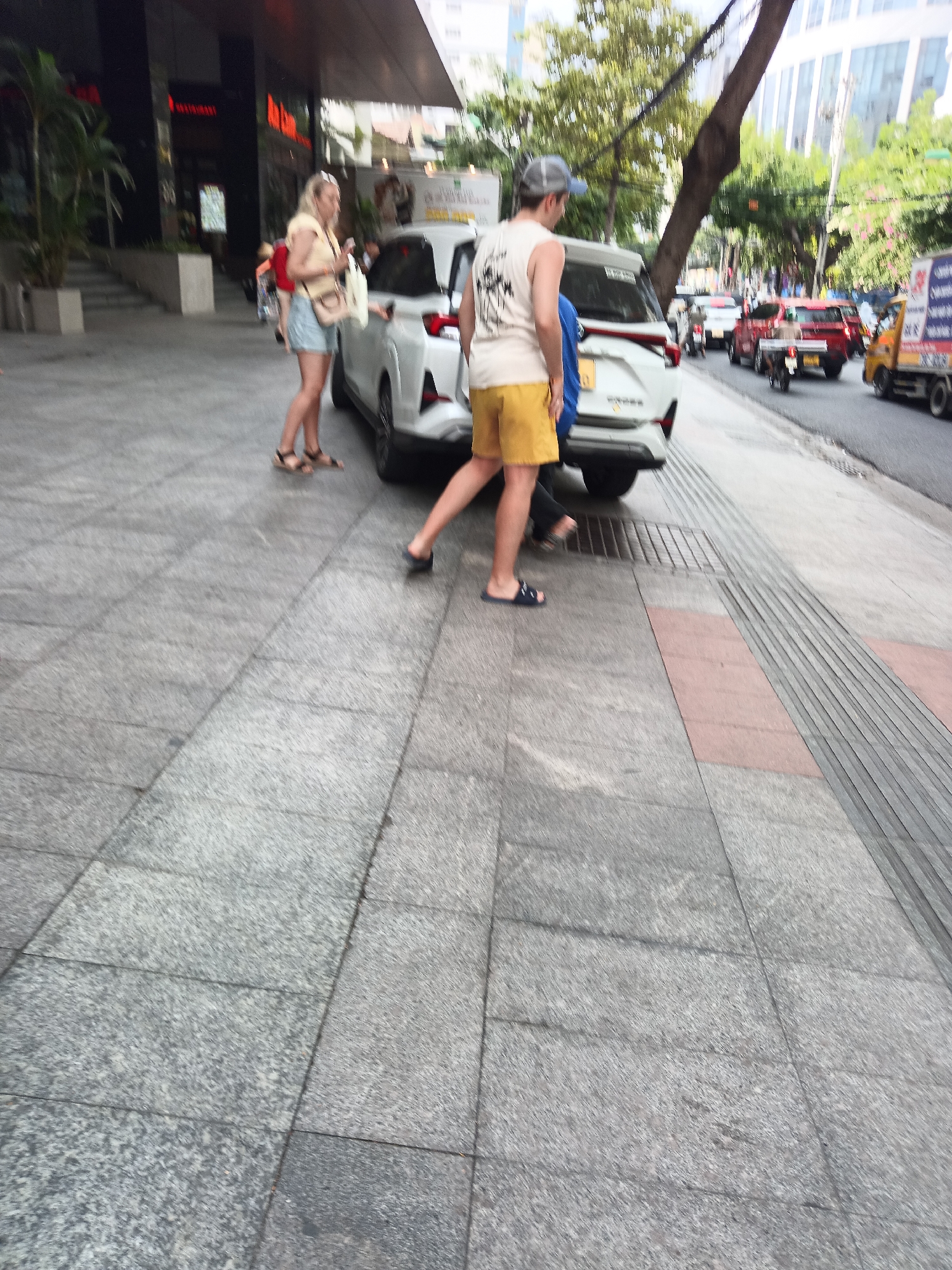 In Nha Trang, Vietnam, a street scene unfolds on a pedestrian walkway paved with large, gray granite tiles. The walkway is bordered by a textured tactile paving strip, designed to assist visually impaired individuals, and a road with ongoing traffic.
In the foreground, a person wearing dark blue flip-flops and yellow shorts walks away from the camera. To their left, a white SUV is parked, with its rear taillights illuminated.
Adjacent to the SUV, a woman in a light-colored tank top and denim shorts stands with her left foot resting on the vehicle's bumper. She is holding a light-colored bag and wearing black sandals. Her hair is blonde and tied back. Another person, partially obscured by the SUV, is visible behind the woman.
In the mid-ground, traffic fills the street. A red car with its brake lights on is stopped, and behind it, a motorcycle carrying a rider is visible. Further down the road, a yellow truck with a blue and white decal on its side is moving. Trees with green foliage line the street, and in the background, modern buildings with reflective windows dominate the skyline. The overall impression is a bustling urban environment with both pedestrian and vehicular activity.