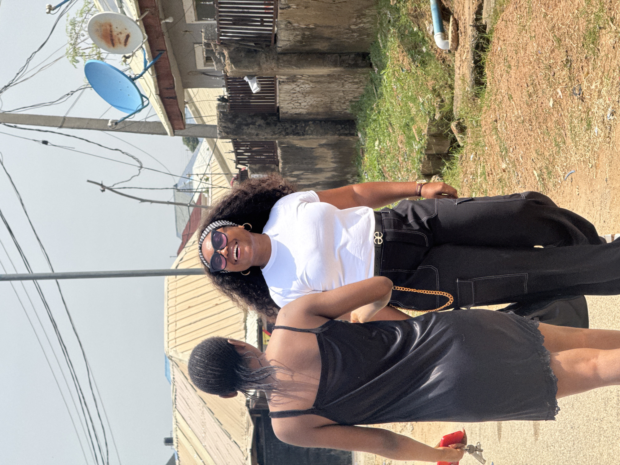 Two women are captured in a candid moment outdoors, likely in an urban environment with residential buildings in the background. One woman, in the foreground and facing away from the camera, wears a black slip dress. The other woman, in the background and facing the camera, is laughing joyfully. She is wearing a white t-shirt, black pants, sunglasses, and a headband. The scene appears to be set during the daytime, with bright sunlight casting shadows. The setting includes elements like electrical wires, satellite dishes, and a grassy, unkempt area, suggesting a developing or less-maintained part of a city, consistent with the provided location context of Abuja, Nigeria. The overall mood is lighthearted and spontaneous. No visible text is present in the image.