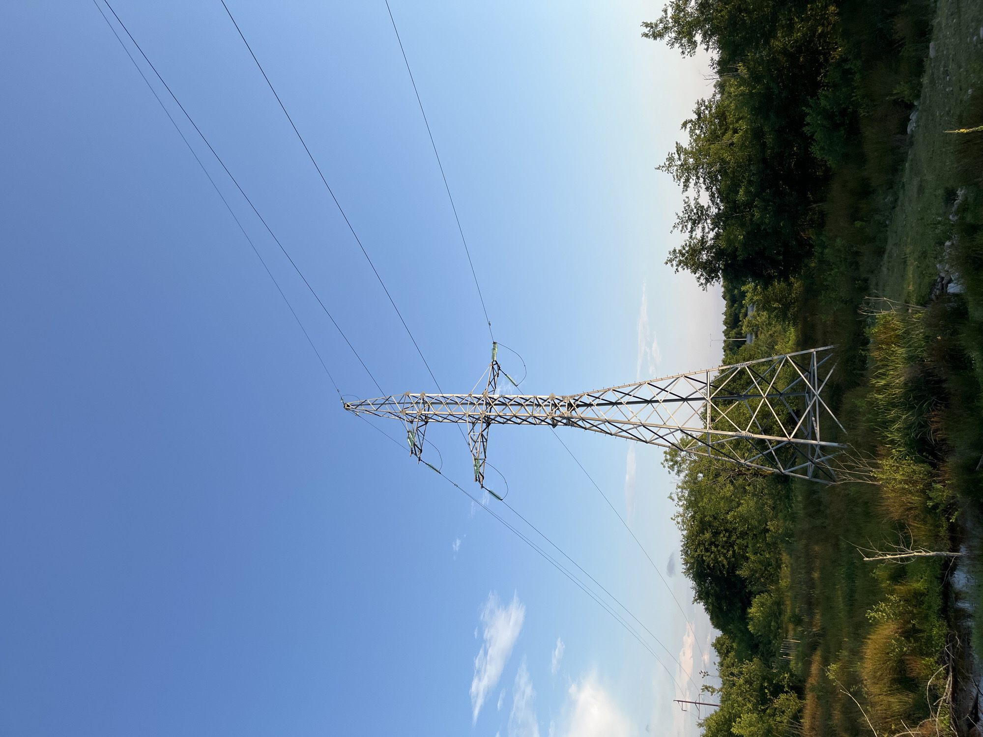 The image depicts a tall, lattice metal electricity pylon situated in a natural landscape. The pylon is centrally located and dominates the frame, with power lines extending horizontally across the clear blue sky. The surrounding environment is lush and green, characterized by dense trees and vegetation, with a body of water or marshy area visible in the foreground. The scene appears to be a rural or semi-rural area in Ureki, Georgia, given the context provided. There are no people or animals present in the image, and the focus is on the industrial structure within its natural setting.