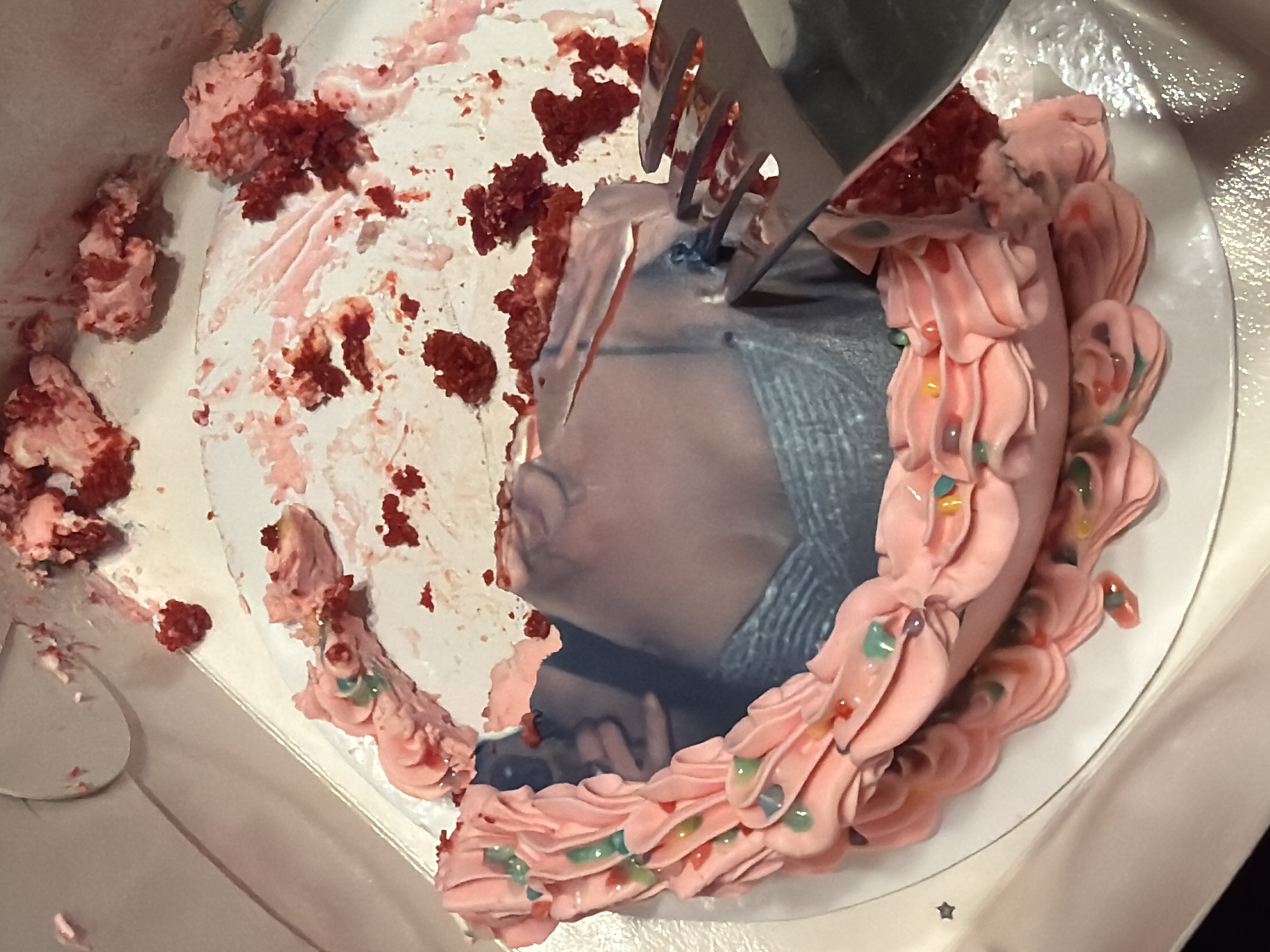 The image shows a partially eaten red velvet cake in a white cardboard box. A metal serving fork is piercing the cake, which has a printed image on the remaining portion. Pink frosting borders the intact part of the cake, adorned with small, colorful candies. Crumbs and smeared frosting are scattered around the eaten section. No information about the location in Belgrade, Serbia, is discernible from the image.