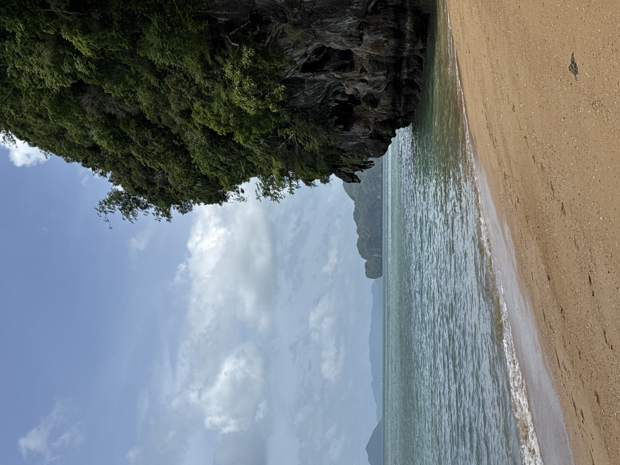 The image shows a tropical beach scene in Khlong Khian, Thailand. On the right side of the frame is a large, sheer cliff face covered in dense green vegetation. The cliff extends into the turquoise ocean, where gentle waves lap against its base. In the foreground, a sandy beach stretches from the bottom right corner towards the left, meeting the water with a thin line of white foam. In the distance, several other islands and landmasses, also covered in lush greenery, can be seen across the calm, blue-green sea. The sky is partly cloudy, with patches of blue interspersed with white cumulus clouds. The overall scene depicts a serene and natural coastal landscape.