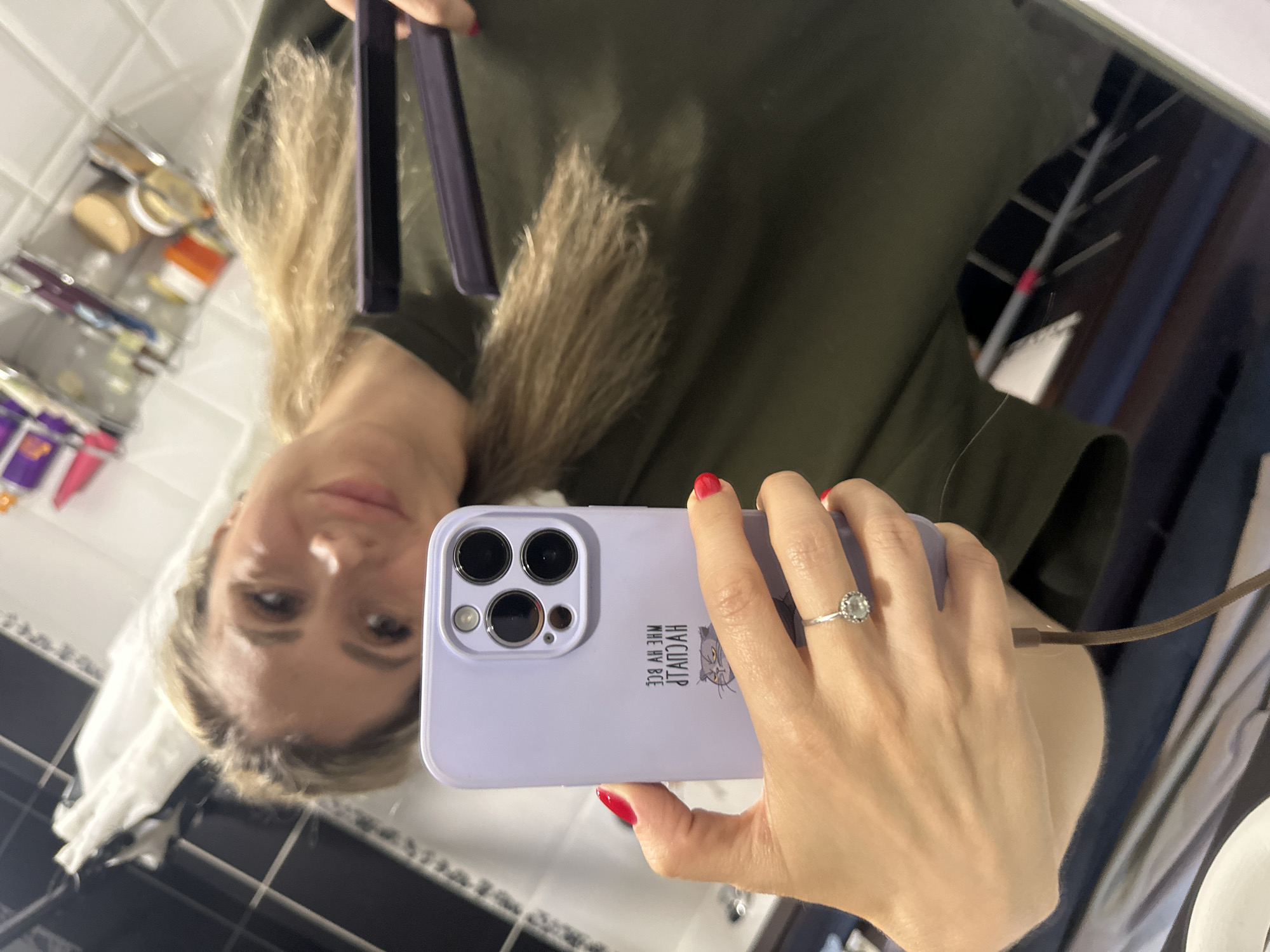 In a dimly lit bathroom with tiled walls, a woman is taking a selfie in a mirror. She has blonde hair and is wearing a green shirt. Her left hand is holding a light purple phone, and her right hand is holding a hair straightener, which she is using to style her hair. A ring with a diamond is visible on her left ring finger. The phone case has a cat design and text in Russian. The bathroom shelves in the background are filled with various toiletries. The image is a reflection captured in a mirror, suggesting the woman is standing in front of it. There is no visible indication of the city of Novoaltaysk, Russia in the image.