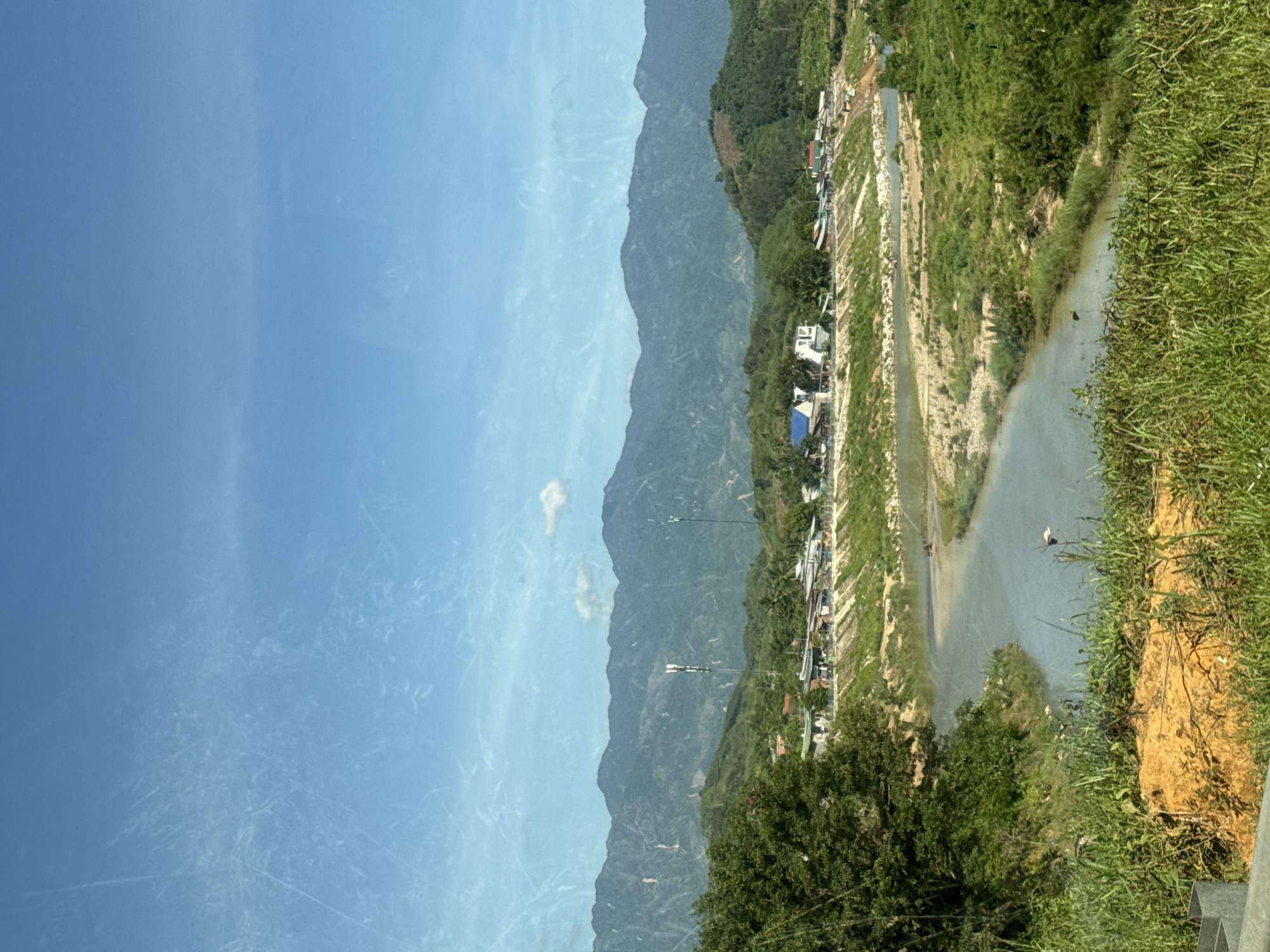 The image depicts a panoramic view of a rural landscape, likely in Nam Khánh Vĩnh, Vietnam, under a clear blue sky with some faint streaks of clouds. The foreground is dominated by tall green grass and a patch of orange-brown soil, suggesting an elevated viewpoint. A winding river meanders through the middle ground, with sandy banks and patches of vegetation. On the far bank of the river, a cluster of buildings, appearing to be houses and small structures, is visible along a sloped embankment, with a road or path running in front of them. Beyond the settlement, lush green hills rise, transitioning into a series of imposing, mountainous peaks in the background. Two tall communication towers are prominently positioned amidst the hills. The overall impression is one of a serene, undeveloped natural environment integrated with a small human settlement. There are no visible people in the image.