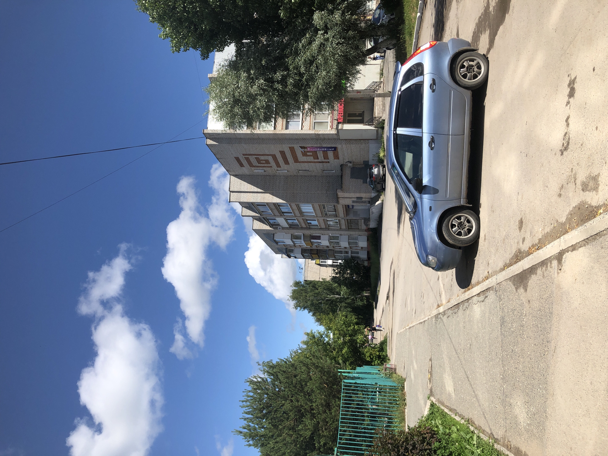 A blue hatchback car is parked on the right side of a paved road in front of a multi-story brick building. A sidewalk runs along the left side of the road, bordered by a green metal fence and trees. The sky above is blue with scattered white clouds. The building appears to be an apartment complex with shops on the ground floor, as indicated by signage. Two people are walking on the sidewalk in the distance. The scene is likely in Solikamsk, Russia, a city known for its industrial and residential areas. The image captures a typical urban street scene on a sunny day.