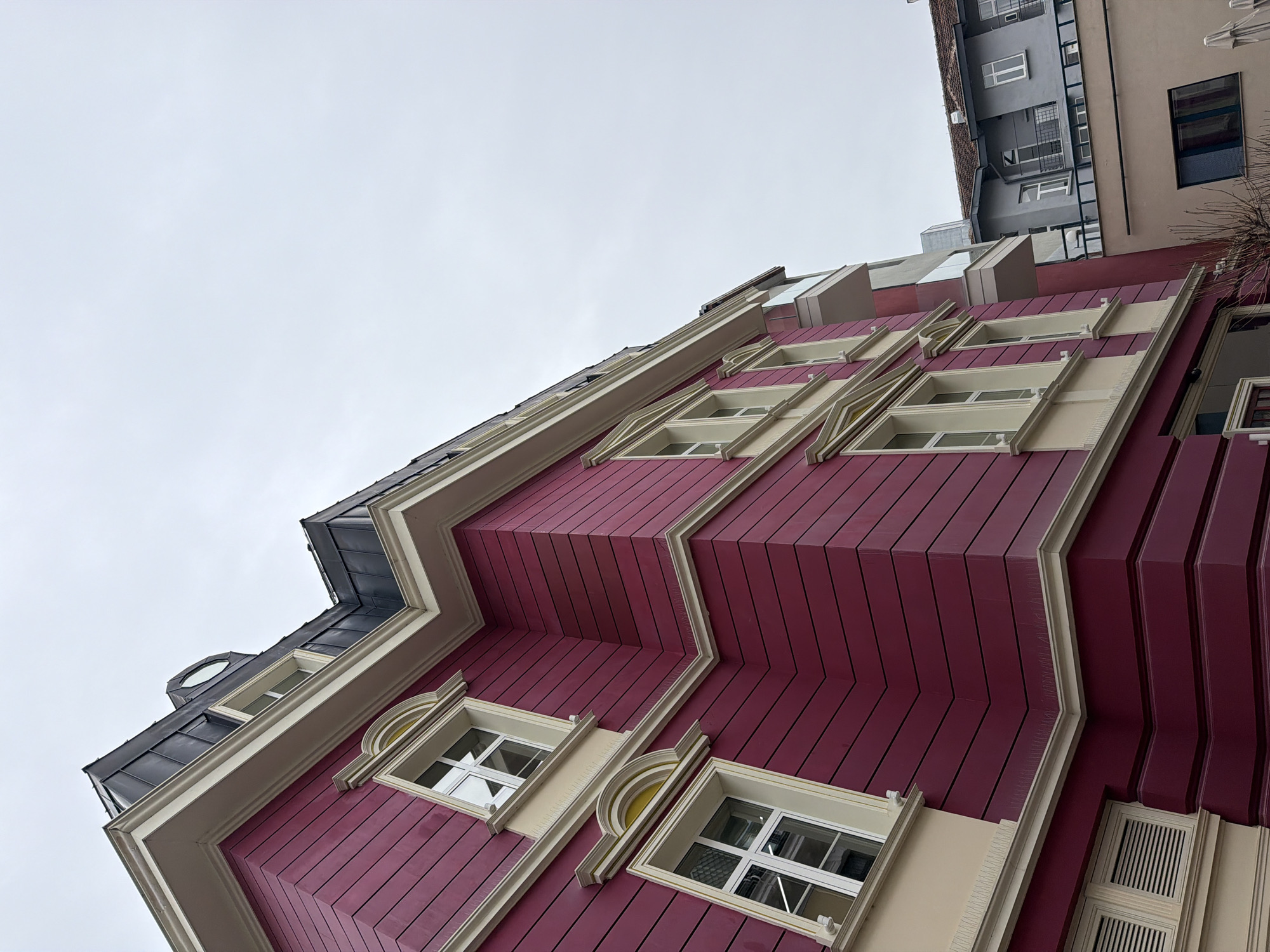 The photograph depicts a building with a distinctive facade, captured from a low-angle perspective. The building features a dark red exterior with light beige trim, and its design incorporates horizontal paneling and decorative elements around the windows. The architectural style appears to be contemporary, with unique, slightly geometric exterior angles.
Above the depicted building, a glimpse of another building of a different style is visible, indicating an urban setting. The sky is overcast, creating a subdued natural light. The image captures architectural detail without any identifiable people or subjects, focusing on the urban environment.
The location is Sofia, Bulgaria.