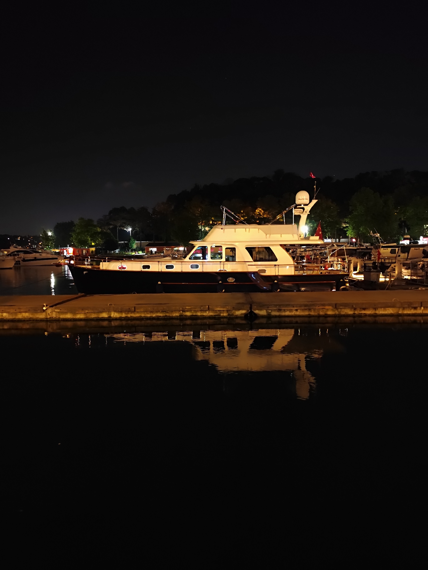 At night, a motor yacht is docked at a pier in Sarıyer, Turkey, likely along the Bosphorus strait. The yacht, predominantly white with a dark blue hull, has several windows and a radar dome on its upper deck. A Turkish flag is visible on the yacht's mast. The pier itself is concrete, with mooring bollards. The water in front of the pier reflects the lights of the yacht and the surrounding environment. In the background, a treeline is illuminated by distant lights, suggesting an urban or suburban area. Other vessels are visible further down the pier, some appearing to be smaller boats or more yachts. The sky is dark, indicating nighttime.