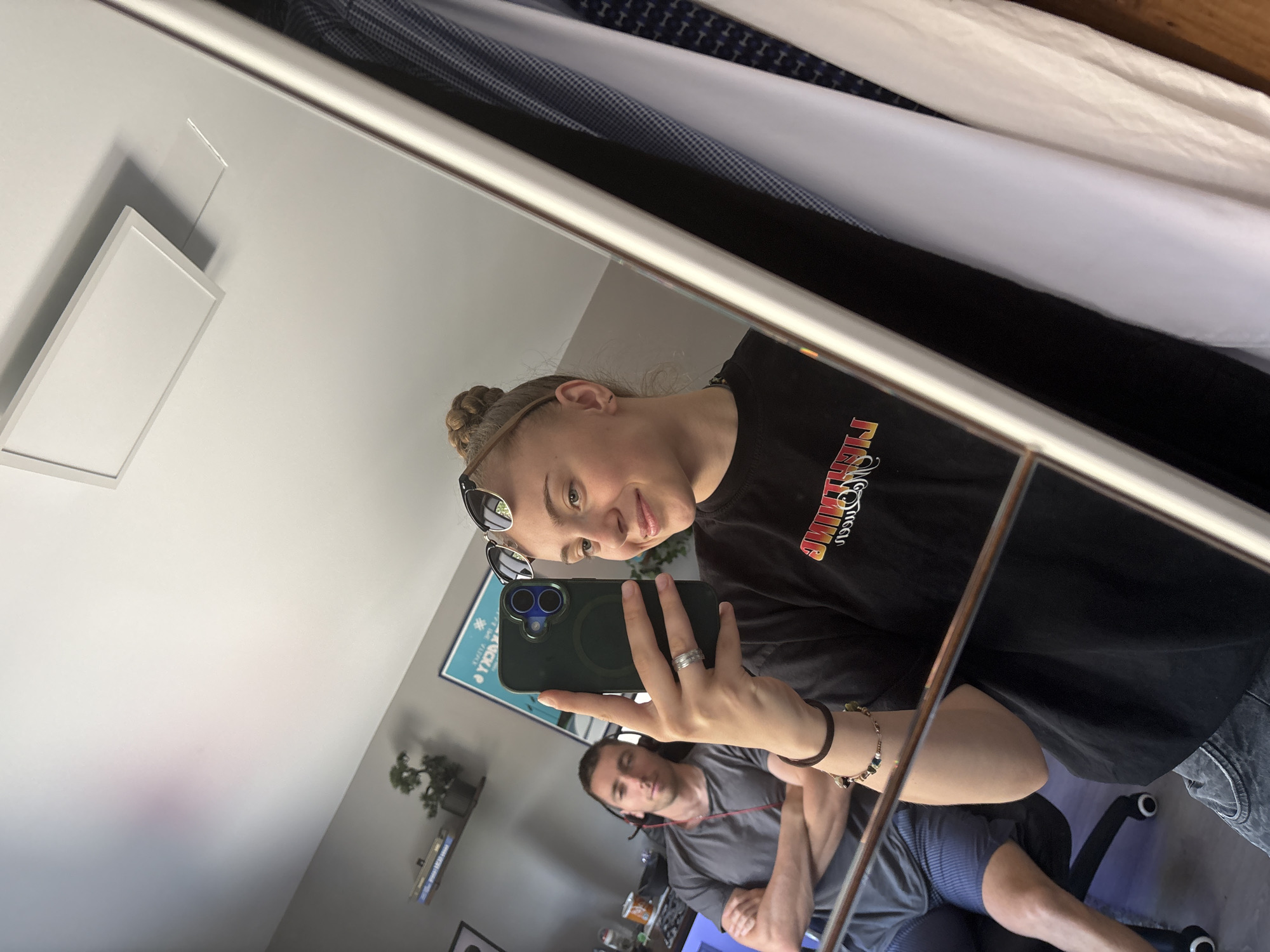 In a selfie taken in what appears to be an apartment in Gabrovo, Bulgaria, a young woman with blonde hair styled in a bun is holding a dark green smartphone up to a mirror. She is wearing a black t-shirt with a colorful graphic and sunglasses perched on her head. Her left hand, adorned with rings and a bracelet, is visible as she holds the phone. Her facial expression suggests she is smiling.
Reflected in the mirror, a man in the background is seated in an office chair, wearing a grey t-shirt and shorts. He has headphones around his neck and his arms are crossed. The room's background includes a desk with a computer monitor, shelves with books and a small plant, and a poster on the wall. The mirror itself reflects a clothing rack filled with garments, indicating the presence of a closet or wardrobe nearby. The overall lighting suggests it is daytime, with natural light entering the room.