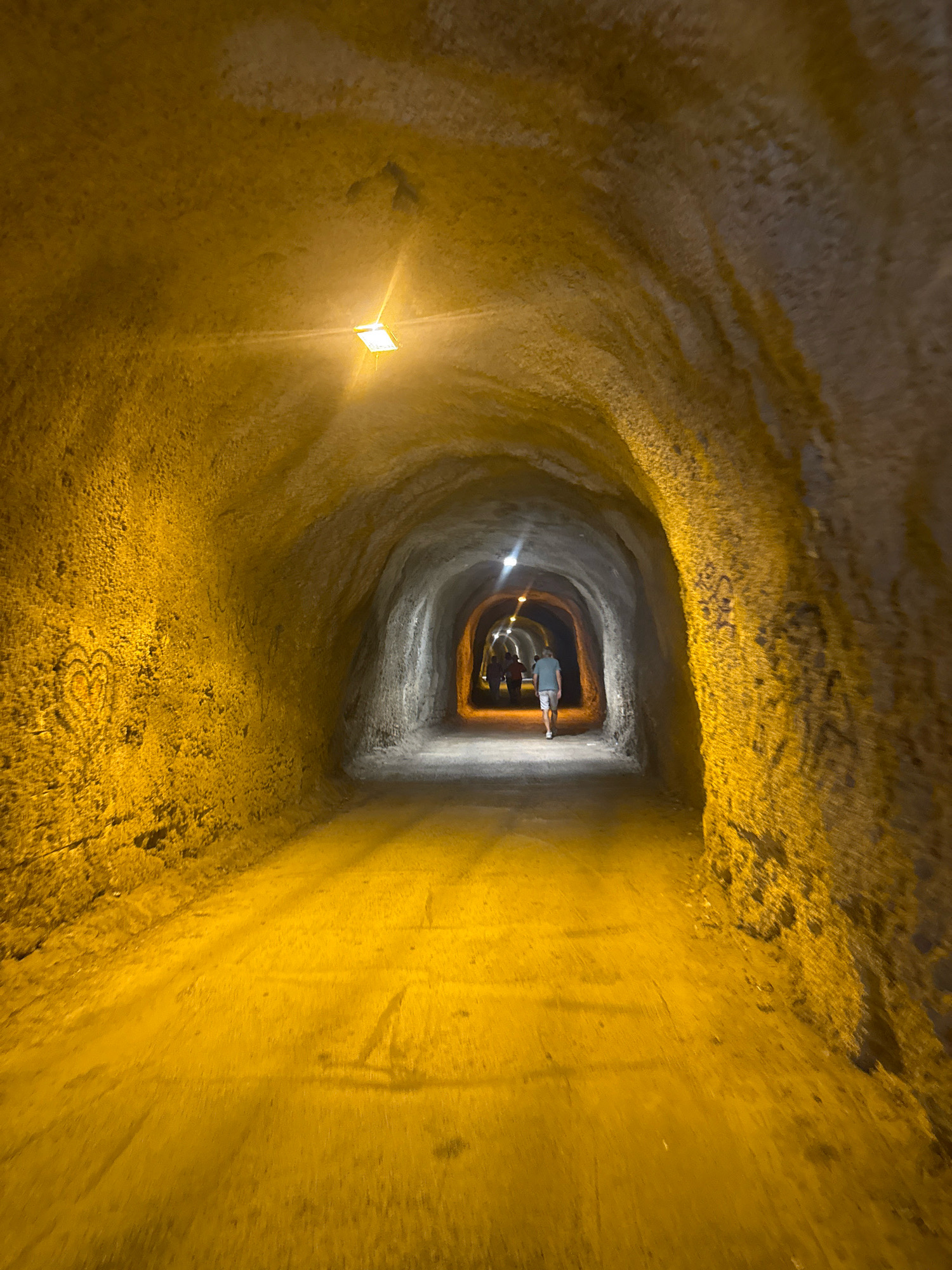 The image shows a tunnel with rough, textured walls, illuminated by yellow artificial lights. The tunnel appears to be made of stone or concrete, with some areas exhibiting darker, possibly damp patches. Faint etchings of a heart shape are visible on the left wall. In the distance, several people are walking away from the camera, entering the darker part of the tunnel. One person in the foreground, facing away, is wearing a light-colored t-shirt and shorts. The tunnel floor is uneven and appears to be a dirt or gravel path. The overall scene suggests an atmospheric and somewhat aged underground passage, possibly a historical or natural formation within the city of Kamenovo, Montenegro.