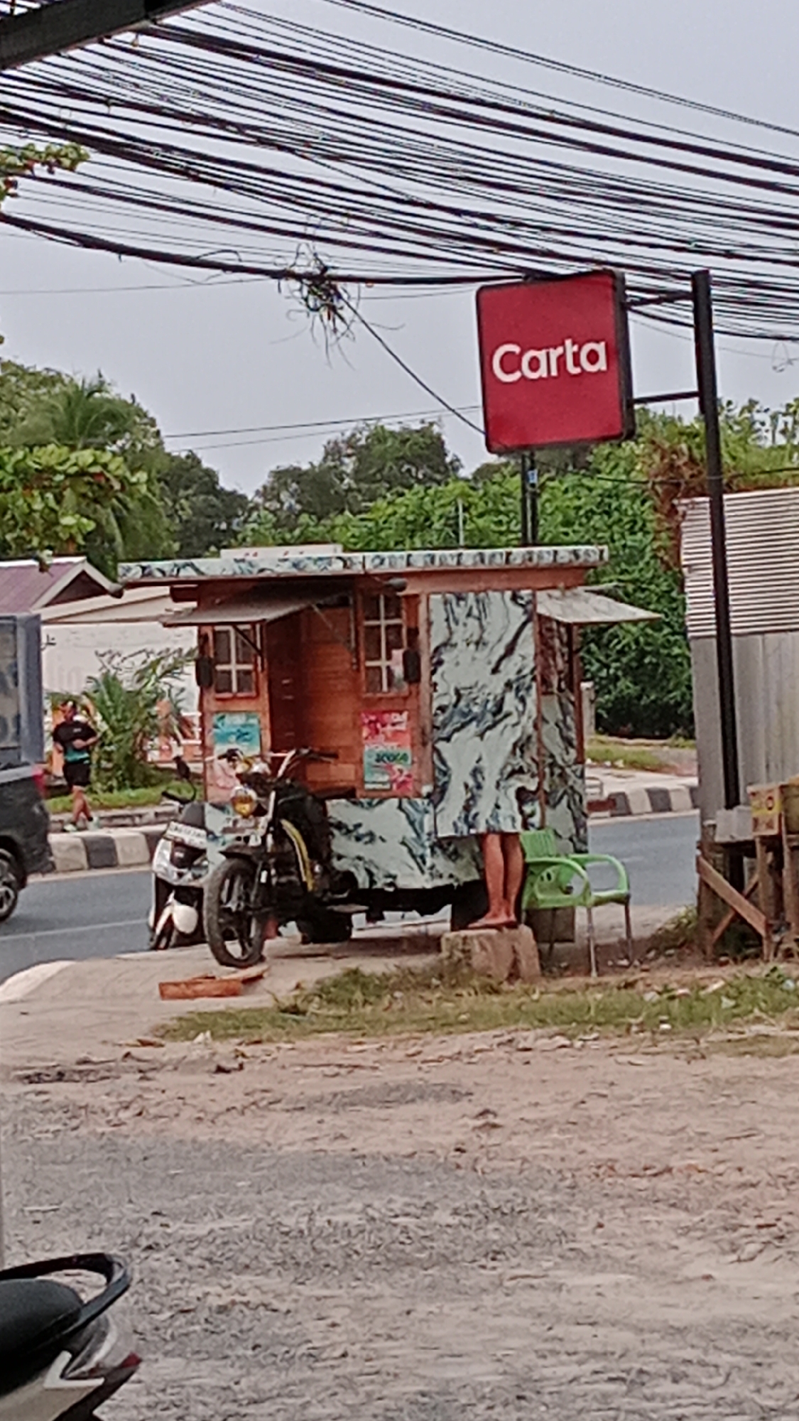In Handil Bakti, Indonesia, a makeshift food stall is set up next to a road. The stall itself is constructed from wood and features a patterned exterior. In front of the stall are two motorcycles, one white and one black. A person's legs are visible behind a portion of the stall, standing on a concrete block. A green plastic chair is positioned near the stall. Above the scene, a multitude of overhead wires are suspended. A red square sign with the white text "Carta" is attached to a pole to the right of the stall. Trees and buildings are visible in the background.