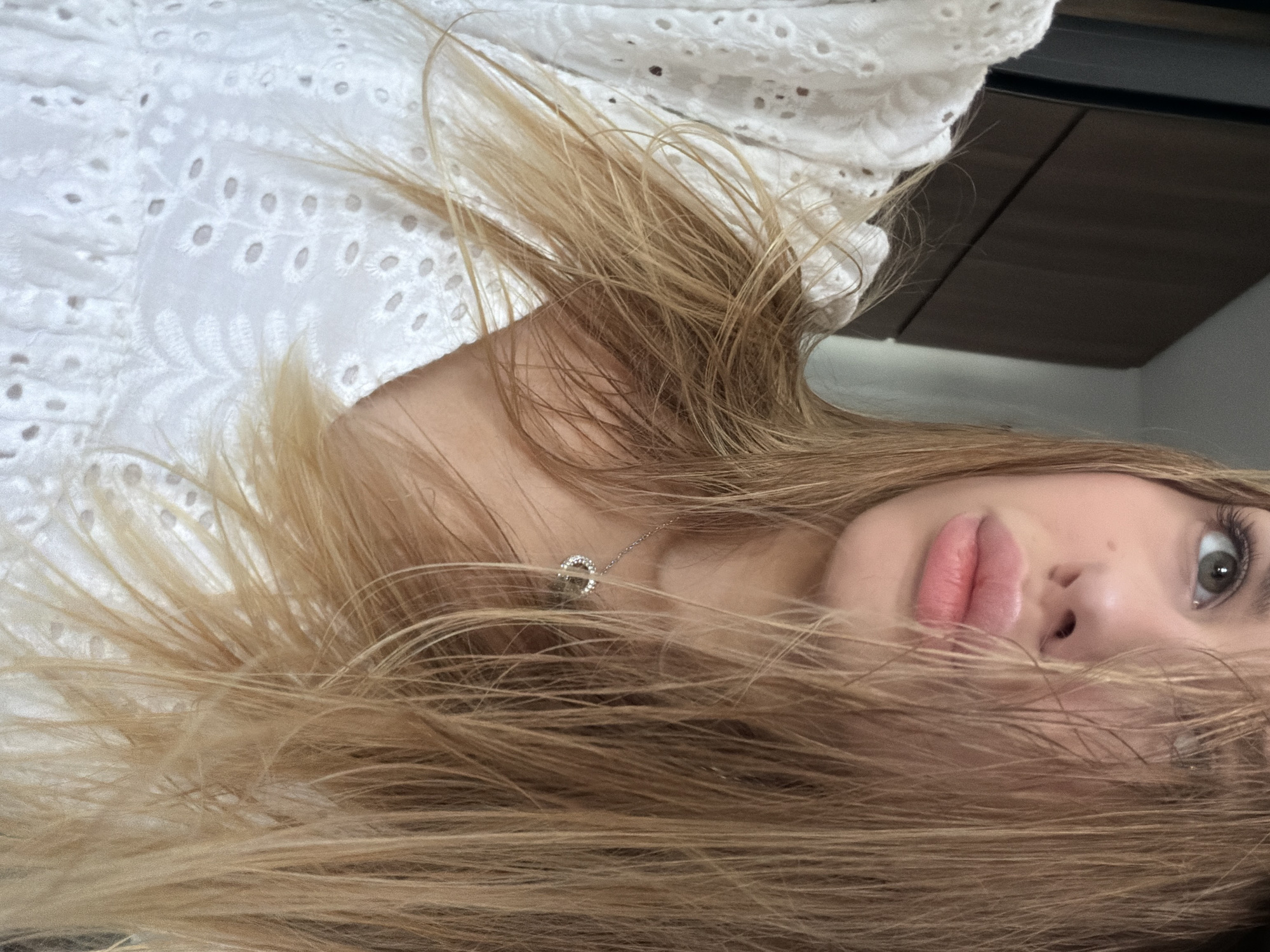 This is a close-up, eye-level, portrait-style photograph of a young Caucasian female. She has long, blonde hair that partially obscures her face. She is wearing a white, eyelet fabric top with ruffles around the neckline and a delicate silver pendant necklace. Her lips are slightly parted, and her gaze is directed upwards and to her left. The background is out of focus and appears to be an indoor setting with dark wooden cabinetry on the left and a lighter colored wall or object on the right. There is no visible indication of the city of Đà Nẵng, Vietnam.