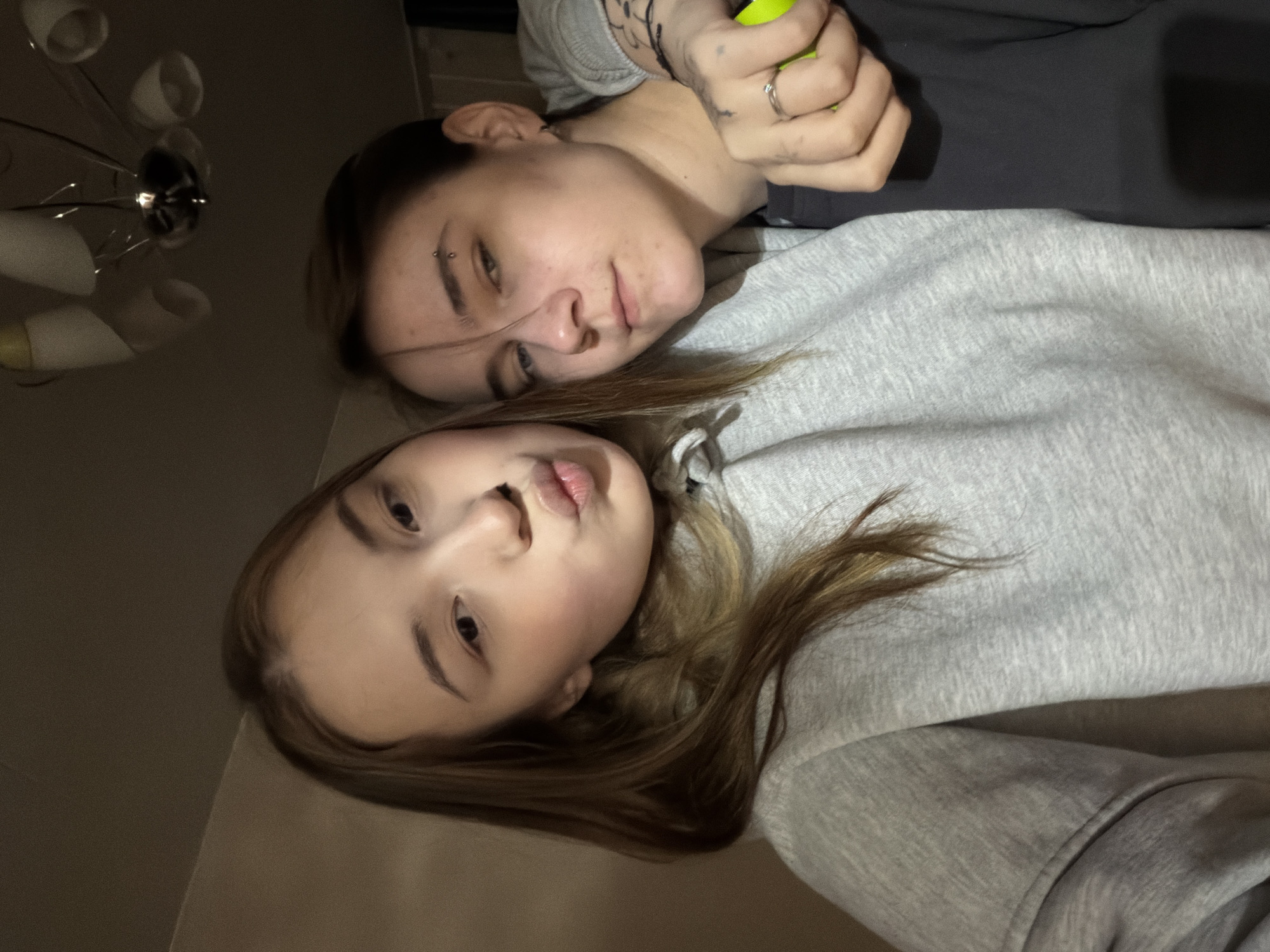Two individuals are visible in the image. The individual closer to the foreground has light skin, light brown hair, and is wearing a grey hoodie. Their facial expression appears neutral. The second individual, positioned further back, has light skin, dark hair, and an eyebrow piercing. They are wearing a grey shirt. Their hand is visible holding a green object. The lighting is subdued. The location is unknown, and there is no indication of Chaikovsky, Russia.