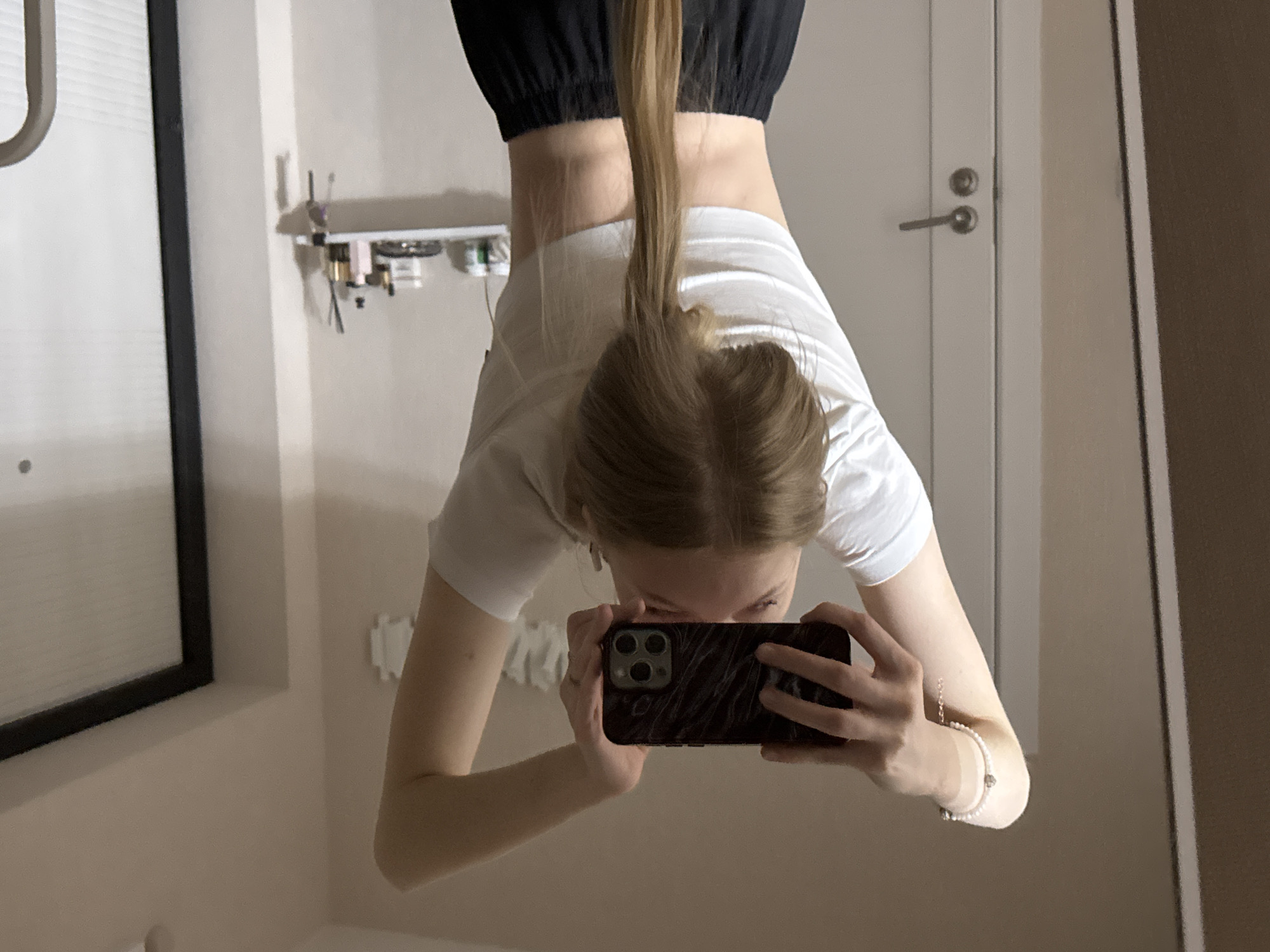 An indoor mirror selfie captures a young individual, likely female, in an inverted position while taking a photo. The subject is seen from the chest up, hanging upside down, with her light brown hair tied in a ponytail cascading towards the top of the image frame. She is dressed in a white short-sleeved t-shirt and black pants. Her face is partially obscured by a dark-colored smartphone, possibly an iPhone due to its triple camera array, which she holds with both hands to capture her reflection. A white beaded bracelet with a charm adorns her left wrist, and a small ring is visible on her right hand.
The reflection reveals a neutral-toned room. To the right of the subject, a light-colored interior door with a silver handle and a round lock is visible. On the left, a small white shelf is mounted on the wall, holding several small items, possibly cosmetics or personal care products, some with thin applicators or brushes extending upwards. Further left, a portion of what appears to be a window with blinds or a framed object is reflected. The overall lighting is soft, suggesting an indoor setting, possibly under artificial light. The image captures a playful or athletic moment, showcasing an unusual inverted perspective. No visible text is present. The scene is located in Perm, Russia.