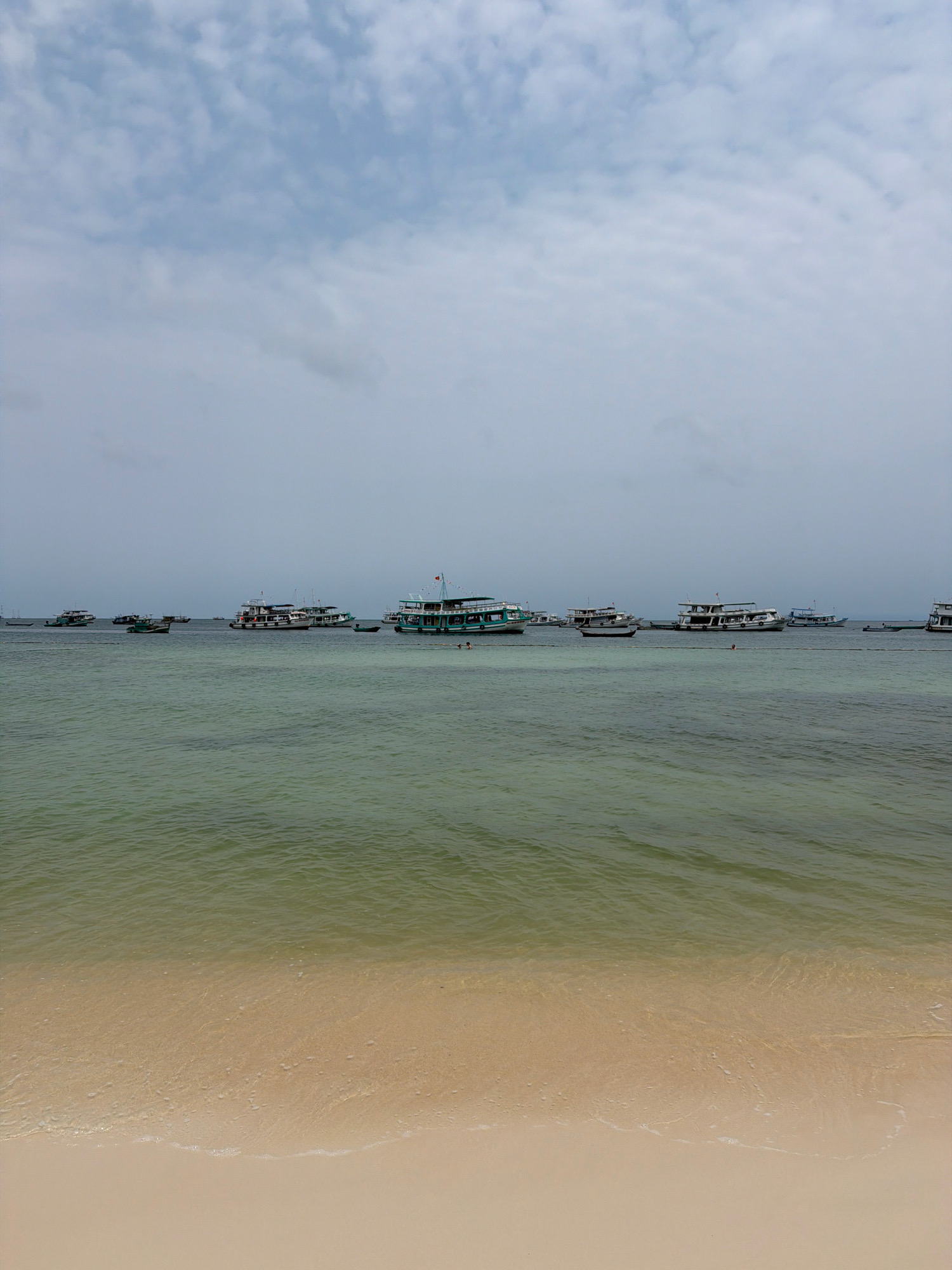 The image captures a serene tropical seascape in Phú Quốc, Vietnam. The foreground features a pristine, sandy beach with gentle waves lapping at the shore. Beyond the beach, the turquoise waters of the ocean stretch towards the horizon, dotted with numerous boats. These vessels, ranging from small fishing boats to larger passenger ferries, are moored or cruising in the calm sea. The sky above is partly cloudy, with a mix of white clouds and patches of blue, indicating a pleasant weather day. The overall scene evokes a sense of tranquility and natural beauty, typical of a coastal resort town.
