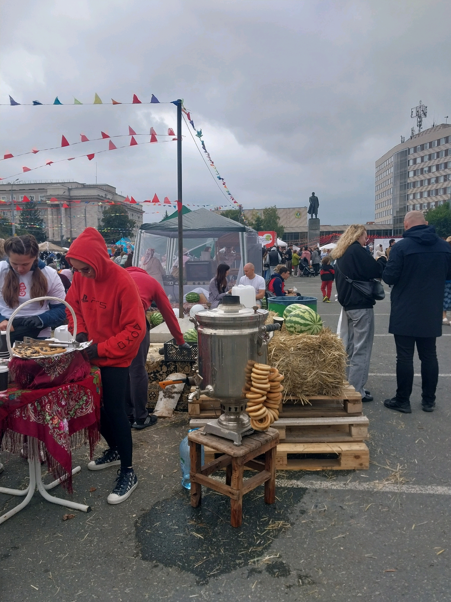 This image captures an outdoor market scene in Orenburg, Russia, with a cloudy sky overhead. In the foreground, a young woman in a red hoodie and black pants is focused on preparing items on a table draped with a red, patterned cloth. Beside her, another woman in a white t-shirt with braided hair is also attending to a basket on the table. A large, traditional samovar stands on a wooden stool, adorned with a stack of round bread rings. Nearby, a large watermelon rests on a bale of hay, placed on wooden pallets. In the background, a crowd of people mill about, browsing stalls and interacting. The setting includes buildings, tents, and festive triangular flags strung across the area, suggesting a public event or fair. The ground is paved and scattered with straw.