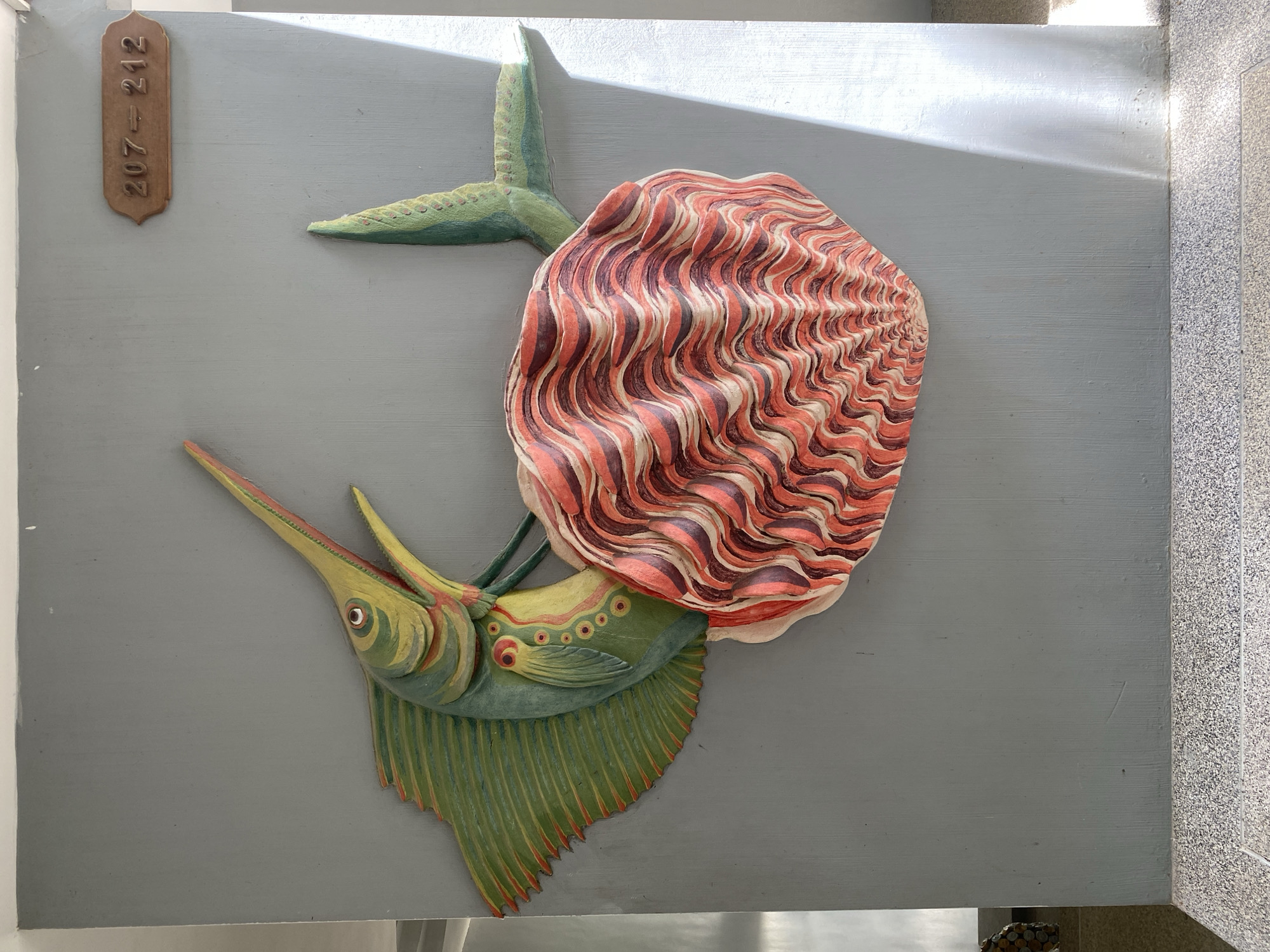 A carved wooden relief of a marlin or swordfish is mounted on a gray wall in a hallway in Koh Phangan, Thailand. The fish, painted in vibrant green and yellow with red accents, appears to be biting a large, intricately carved shell in shades of coral and deep red. The shell is textured to mimic the ridged appearance of a clam or scallop. Above and to the right of the artwork, a rectangular bronze plaque with the numbers "207-212" is affixed to the wall. The wall itself has a subtle textured finish. Sunlight casts a diagonal beam across the right side of the image, illuminating the texture of the wall and creating distinct shadows. The floor below is a speckled terrazzo material.