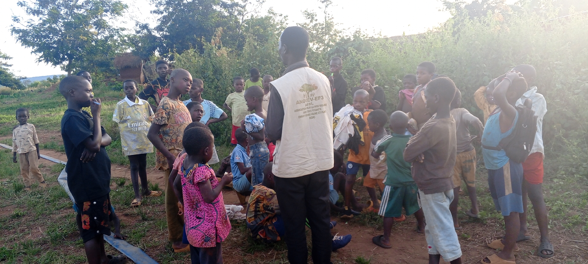 In an outdoor setting in Ngalula, Democratic Republic of the Congo, a group of about 30 children is gathered around an adult male figure. The man is wearing a vest with organizational details inscribed on the back: "ANOVEV-EF3 ASBL ASSOCIATION POUR LA NON VIOLENCE AURI TRES VIVANTS ENFANT-FEMME-FAUNE ET FLARE RDC". The children vary in age, dress, and pose. Some are looking directly at the man, some are looking at other children, and a few are looking away. The background is lush with trees and vegetation. The light is bright with some lens flare. A blue pipe runs through the foreground.