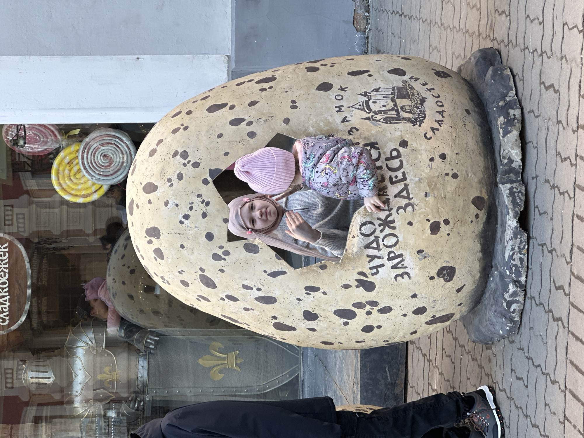 A low-angle outdoor shot captures two young girls within a large, egg-shaped sculpture, standing on a paved sidewalk. The sculpture is beige, adorned with brown speckles and inscribed with Cyrillic text and a castle drawing. The girls are positioned inside a square opening in the egg, with one girl wearing a pink beanie and the other a headscarf. Their attire includes a grey sweater and a unicorn-patterned jacket.
The sculpture rests partially on a rough, grey rock base. The background includes a storefront window displaying lollipops and reflections of a medieval knight statue. A sign with the word "сладкоежек" is visible. A portion of a person wearing black pants and black and white sneakers is visible at the bottom of the frame. The location is Kazan, Russia, identifiable by the Russian text on the egg sculpture.
