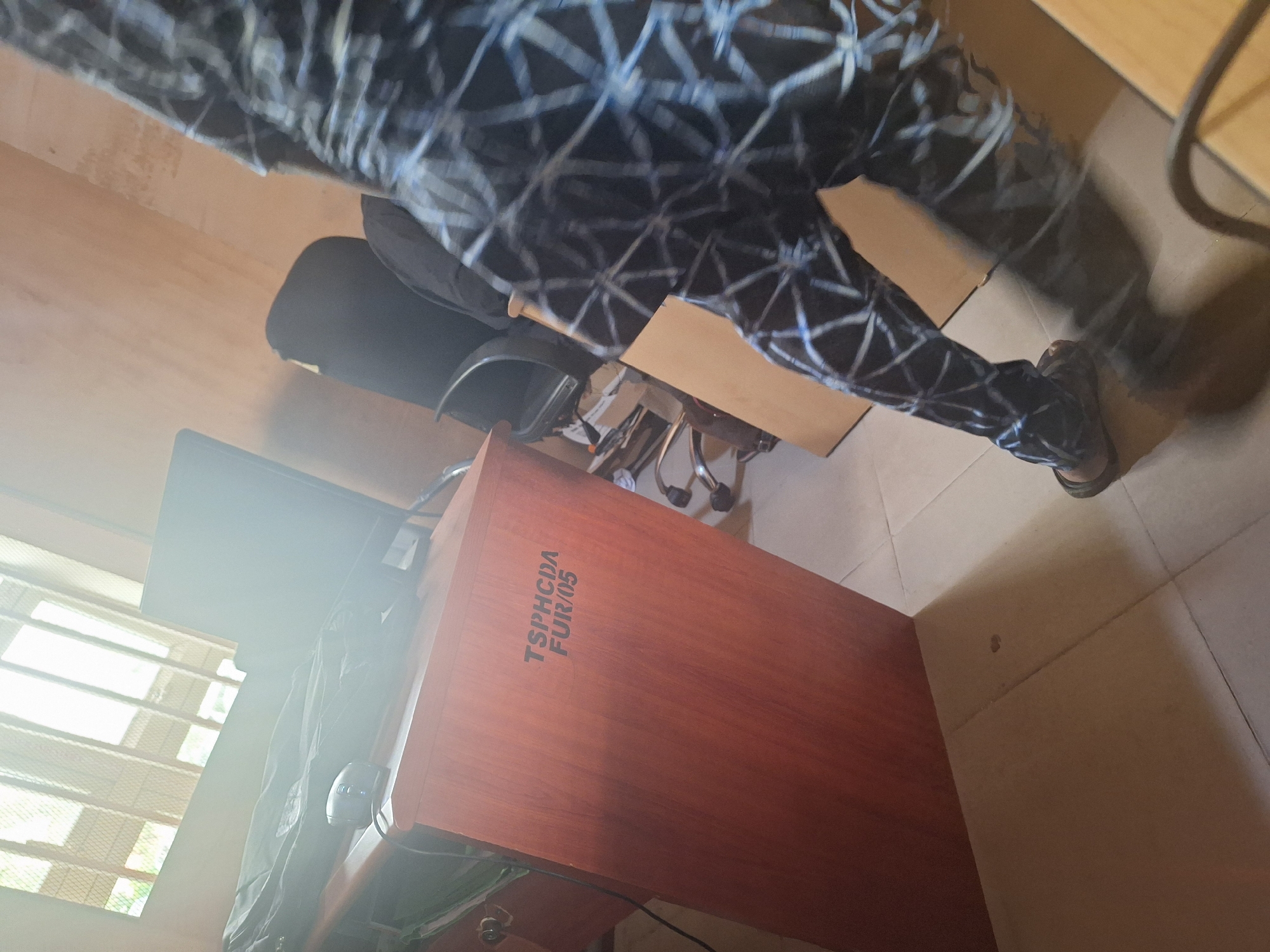 In Jalingo, Nigeria, a person walks in an office. The person wears a patterned shirt and pants, and dark sandals. The person is moving away from a desk and towards the right side of the frame, casting a shadow. The desk is made of wood and has text printed on its side. A computer monitor, a mouse, and a black bag are on the desk. In the background, two office chairs are visible, along with a window covered by blinds. The room has light-colored walls and a tiled floor.