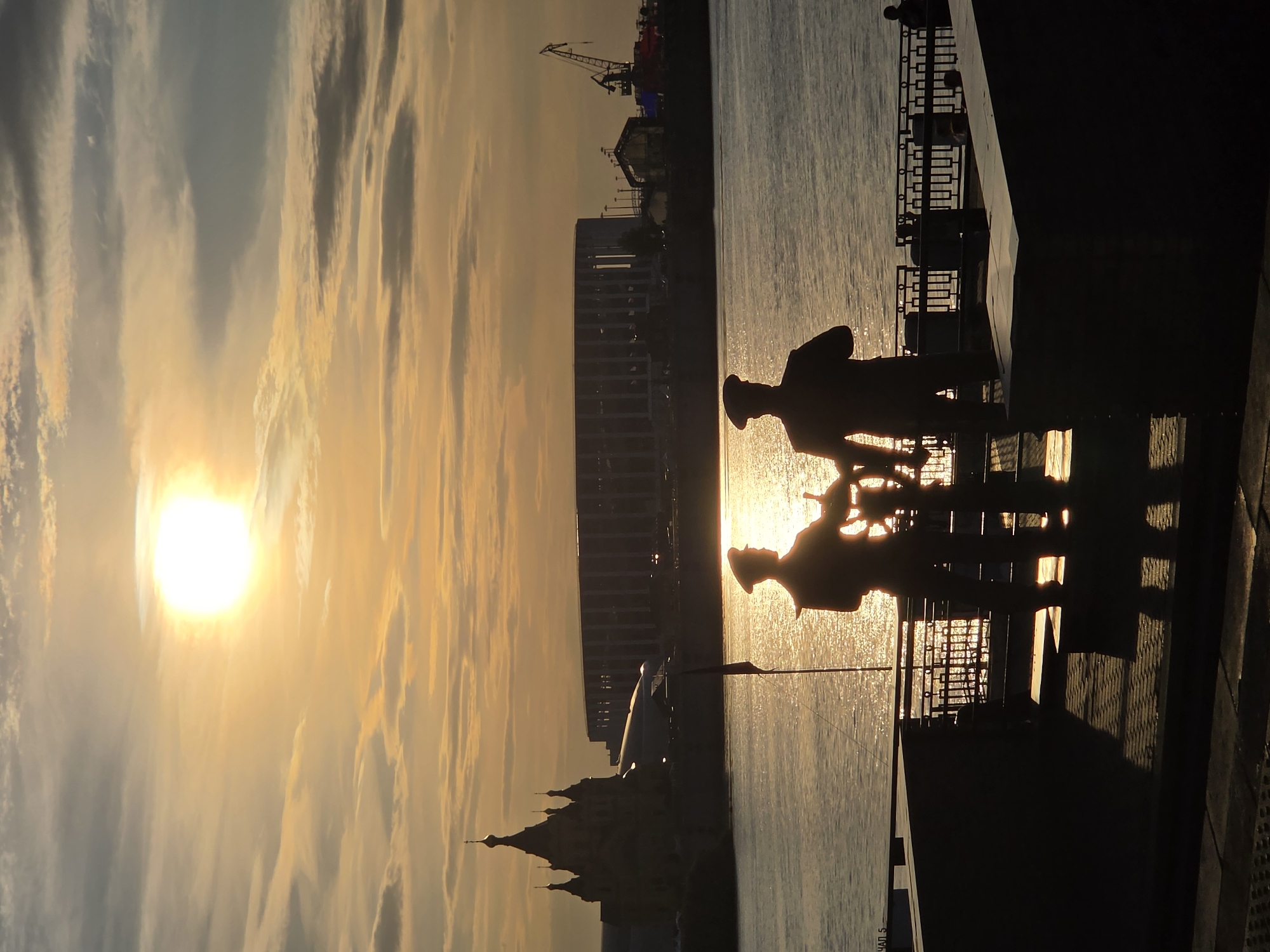 The image depicts a silhouetted scene at sunset on the Nizhny Novgorod embankment, Russia. In the foreground, two statues of sailors stand facing the water, one with his hands on a ship's wheel. Behind them, the sun is low in the sky, casting a golden light across the clouds and the Volga River. The skyline in the distance features a distinctive onion-domed church (likely the Annunciation Cathedral) and a modern stadium. Cranes and industrial structures are visible further along the waterfront. The overall atmosphere is tranquil and picturesque, with the warm light of the setting sun creating dramatic silhouettes.