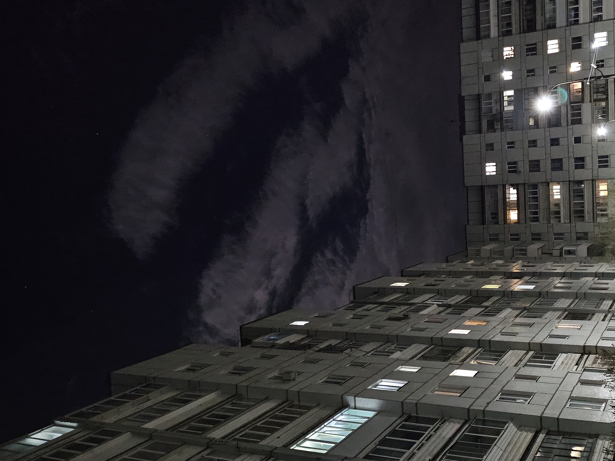 The image presents a nighttime upward perspective of two large, multi-story concrete apartment buildings under a dark, cloudy sky. The architecture is characterized by a brutalist or socialist-era style, featuring numerous rectangular windows across repetitive, unadorned grey facades.
The building on the right occupies a significant portion of the upper right frame, showcasing many illuminated windows with varying light temperatures – some warm yellow, others cool white. A bright streetlight, positioned off-center to the right, emits a strong glow and lens flare, illuminating parts of the building.
The second building originates from the bottom left corner and extends diagonally towards the center, appearing to recede into the distance. Its lower floors, particularly in the foreground, have many visible windows, several of which are brightly lit, casting a soft glow. The perspective emphasizes the vastness and uniform design of the structures.
The sky is a deep indigo-black, indicating night, with prominent, wispy grey and dark cloud formations stretching across the expanse. A few faint, tiny white points, possibly stars, are discernible in the darkest parts of the sky. No individuals or specific activities are explicitly visible, but the illuminated windows suggest life within the residential units. There is no visible text in the image. The scene conveys an atmosphere of quiet urban scale and density at night.