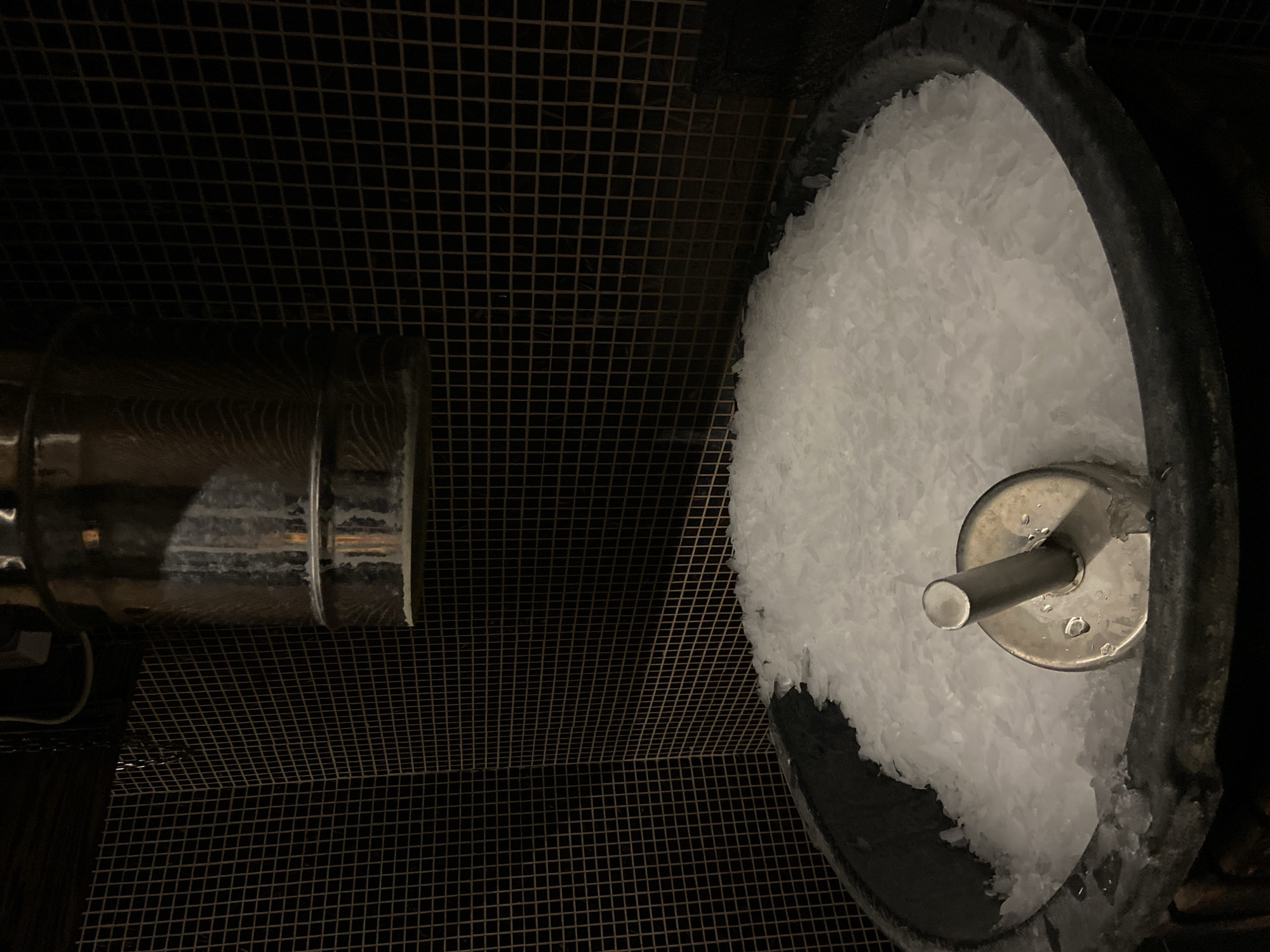The image captures a dimly lit interior scene, likely an industrial or commercial setting such as a kitchen or bar in Kaliningrad, Russia. No people are visible.
On the right side of the frame, a dark, round container or bin is prominently featured, filled with a large quantity of white, flaked or crushed ice. A metallic, cylindrical rod or pipe emerges from the center of the ice, connected to a circular metal plate. Water droplets are visible on this metallic fixture, indicating condensation or melting ice. The rim of the container is dark and appears to be made of a thick, possibly rubberized or plastic material.
To the left, a large, reflective, metallic cylinder or pipe runs horizontally. Its surface shows some light reflections and faint patterns. The background and much of the floor area are covered by a dark, grid-like metal mesh or grating, giving the space an utilitarian appearance. The overall lighting is low, contributing to a stark and functional atmosphere. No text is visible in the image.