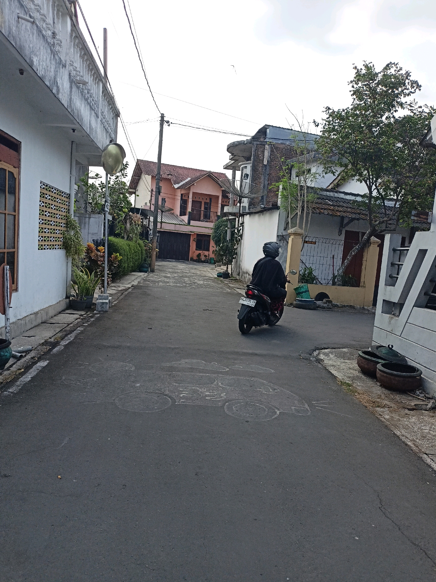 The image depicts a street in Payaman, Indonesia, under an overcast sky. The street is paved with asphalt and shows signs of wear, including chalk drawings of vehicles. On the left side of the street, a white building with decorative architectural elements and a wooden-framed window is visible. Potted plants and a lamppost are situated along the sidewalk. On the right side, a beige building with a brown door and a tiled roof is partially obscured by trees. Further down the street, a light orange house with a dark garage door and a similar tiled roof is present. In the middle of the street, a person wearing dark clothing and a helmet is riding a dark-colored motorcycle. The motorcycle's license plate is visible, reading "AA 2375 TB." The overall environment suggests a residential area.