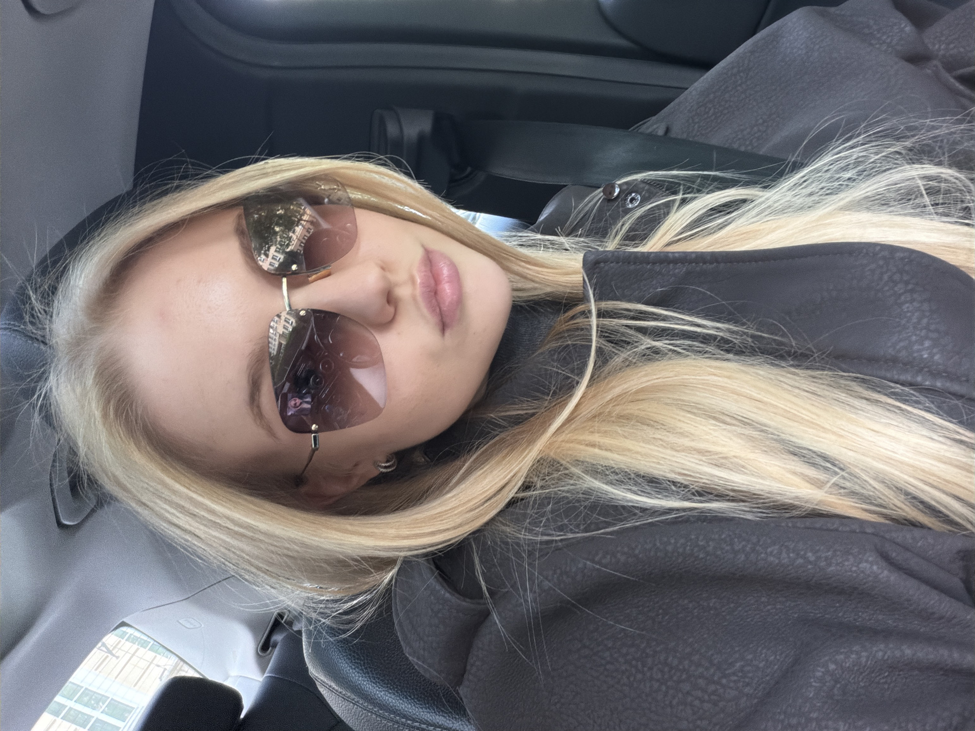 The image shows a fair-skinned young woman with long, blonde hair and sunglasses in what appears to be the passenger seat of a car. The sunglasses reflect a building, suggesting an urban setting. She is wearing a dark jacket. The car's interior is visible, including parts of the seat, dashboard, and possibly a seatbelt. The location is identified as Unknown, Unknown.