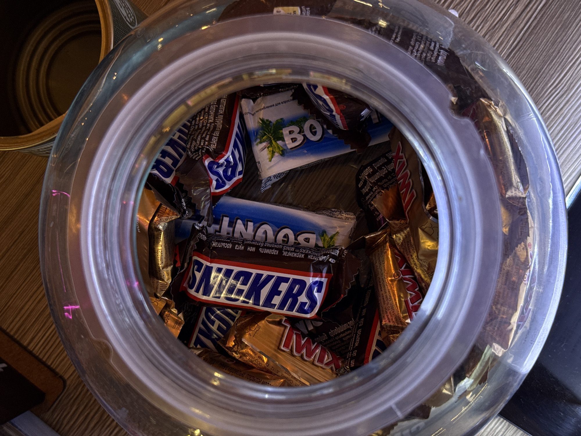 A jar is filled with candy bars. A mixture of Snickers, Twix, and Bounty miniatures are visible. Text on the packaging indicates that the product is distributed by "Mars," with a Moscow, Russia address provided. The candies are encased in their individual wrappers. The jar itself is made of clear glass and has a ridged opening. A can is partially visible near the top left of the image. The jar appears to be sitting on a wooden surface.