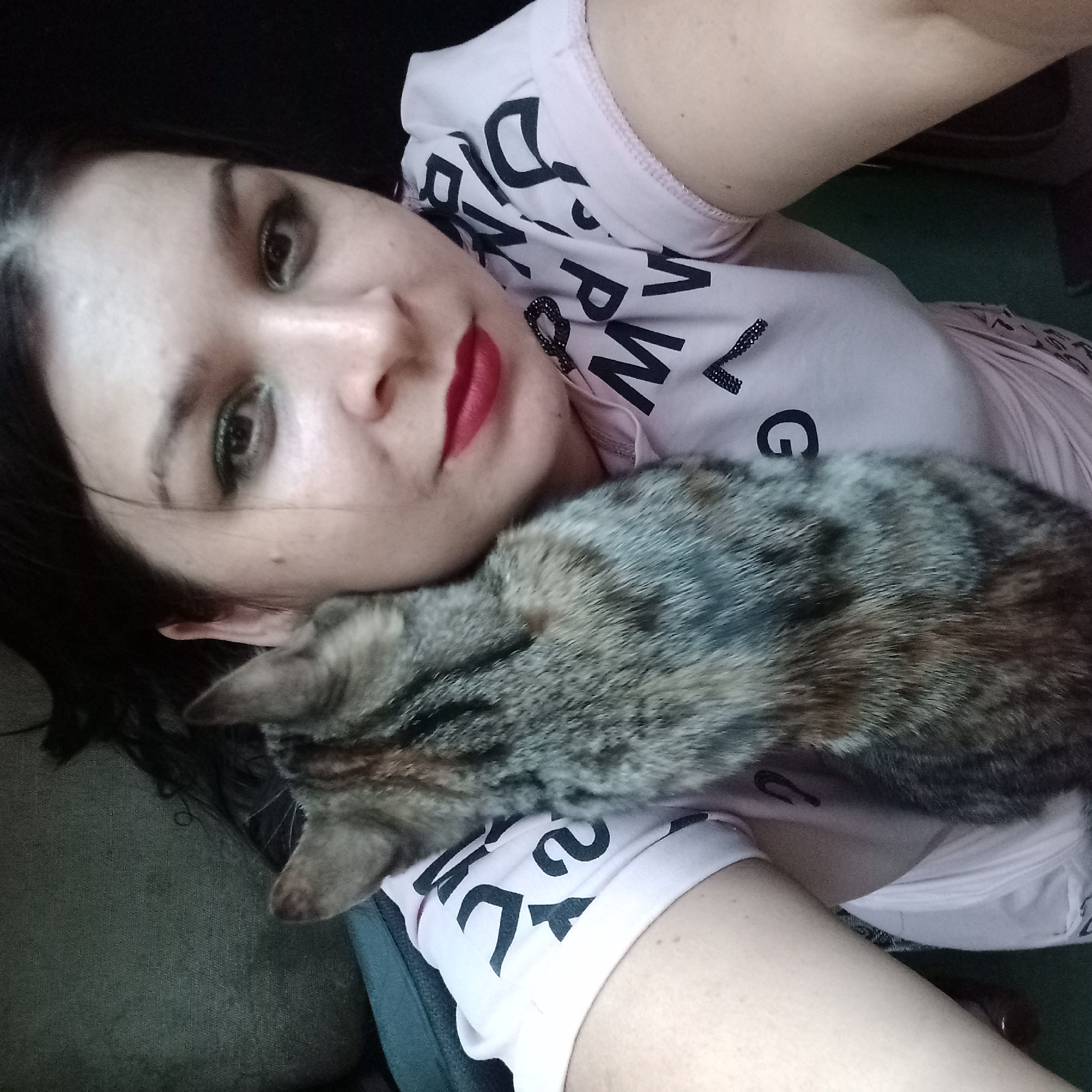 In a close-up, high-angle shot, a woman with dark hair and dramatic eye makeup lies on her back, her head tilted back. She is wearing a light pink t-shirt with black lettering. A tabby cat is lying on her chest and neck, its head resting on her face near her eye. The woman's lips are painted a vibrant red. The background is dark and appears to be indoors, possibly a living room or bedroom.