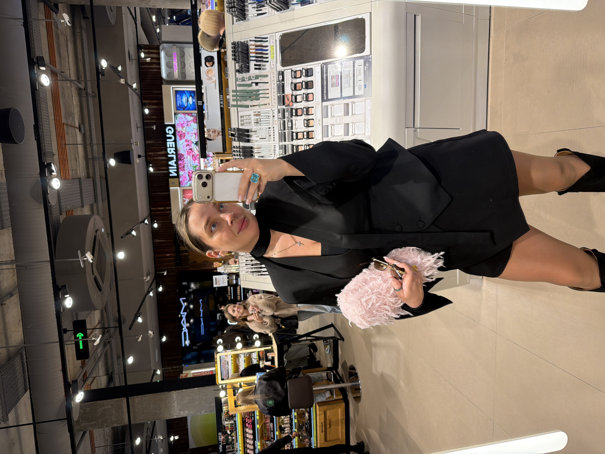 A person is taking a selfie in a store in Moscow, Russia. The individual is holding a smartphone in their left hand, capturing their reflection in a mirror. They are wearing a black dress, a black blazer, a choker, and black shoes. They have a pink fluffy purse and glasses in their right hand. A large blue ring is visible on their left ring finger.
The background is a busy retail environment. In the mirror, there are cosmetic displays, and nearby there are other customers and store personnel. In particular, one customer is seen sitting at a vanity, likely getting a makeover. The overall atmosphere is bright, due to artificial lighting in the store. Text in Russian is visible on displays in the background.