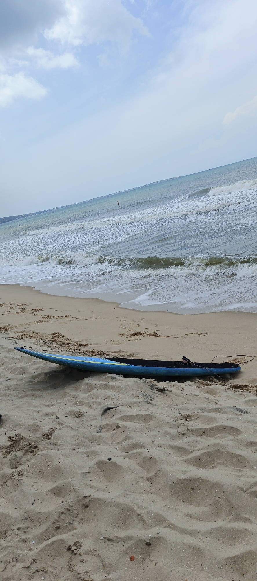 A long blue surfboard with a black leash lies on a sandy beach. The ocean with small waves is visible in the background under a cloudy sky.