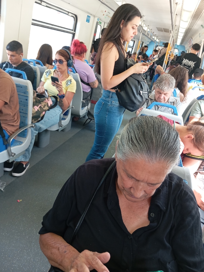 The image depicts the interior of a commuter train car in Castelar, Argentina. The train features a clean, modern design with light blue and grey upholstered seats, white walls, and a textured grey floor. Large windows on the left side of the car illuminate the scene with bright daylight.

In the immediate foreground, an elderly female with grey hair pulled back, wearing a black collared shirt, is seated and looking down, her hands positioned as if interacting with a small object or device. A black strap is visible over her shoulder. Standing in the aisle just behind her, a young adult female with long dark hair, wearing a black tank top and blue jeans, holds and observes a smartphone. She carries a black backpack.

To the left, a female passenger with reddish-brown hair, wearing blue headphones and sunglasses, is seated in a light blue seat. She is looking at a smartphone, dressed in a yellow t-shirt with a green cartoon graphic. Next to her, a young adult male with dark hair, wearing a dark t-shirt and white earbuds, is also seated and looking downwards. Across the aisle on the right, an older male with grey hair, wearing a light-colored shirt, is seated and appears to be resting or looking down. Further back in the car, additional passengers are visible, some standing and some seated, with one individual wearing a black t-shirt with a white graphic. Most visible individuals are engaged with personal electronic devices. The overall atmosphere is that of a typical, busy commuter journey.