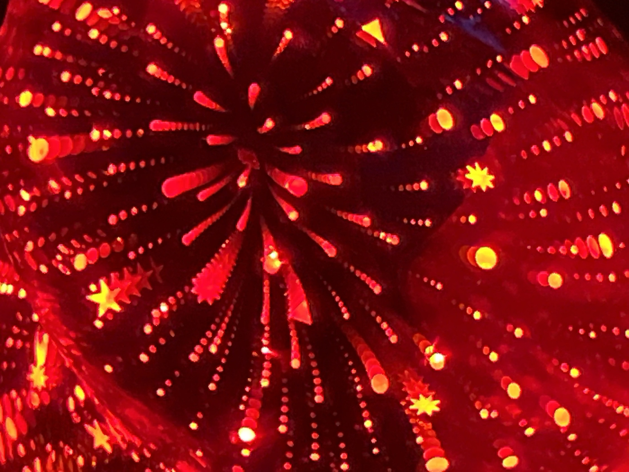 This is a close-up, abstract image of red and yellow lights that appear to be out of focus. The lights are arranged in various patterns, including dots, streaks, and star shapes. Some of the star shapes are sharp and distinct, while others are blurred and indistinct. The overall impression is one of a festive or celebratory atmosphere. The image does not contain any identifiable landmarks, people, or specific objects that would allow for a precise location to be determined. The scene appears to be a light display, possibly from fireworks, holiday decorations, or a light art installation. The colors are predominantly deep red with vibrant yellow highlights.