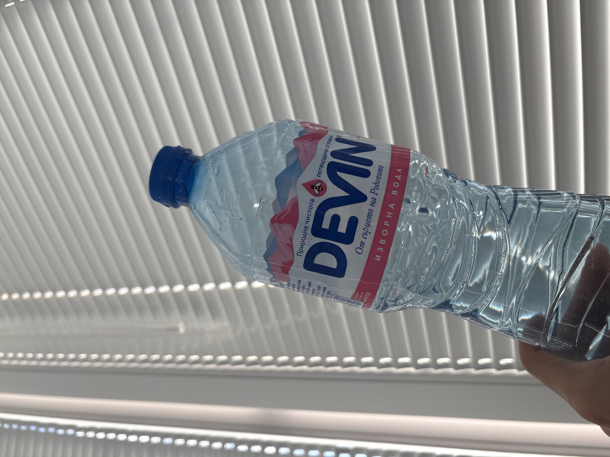 A close-up, angled shot captures a hand holding a clear plastic bottle of "DEVIN" brand spring water. The bottle, nearly full, shows condensation on its surface. The label, designed with white, red, and blue colors, prominently displays "DEVIN" in large blue letters. Below the brand name, the Bulgarian text "От сърцето на Родопите" (From the heart of the Rhodopes) and "ИЗВОРНА ВОДА" (Spring Water) are visible. Above "DEVIN", "Природна чистота" (Natural purity) and "потвърдена от НЕМ" (confirmed by NEM) are printed, along with a small logo featuring a bee or wasp. The bottle is capped with a distinctive blue lid.
The background consists of evenly spaced, light-colored horizontal slats, creating a distinct pattern of bright highlights and shadows. This architectural feature resembles venetian blinds or a slatted ceiling, suggesting an indoor setting with abundant natural light, indicating it is daytime. The hand holding the bottle is visible in the bottom right corner of the frame. The overall impression is clean and well-lit. The presence of Devin water, a well-known Bulgarian brand, confirms the context of Sofia, Bulgaria.