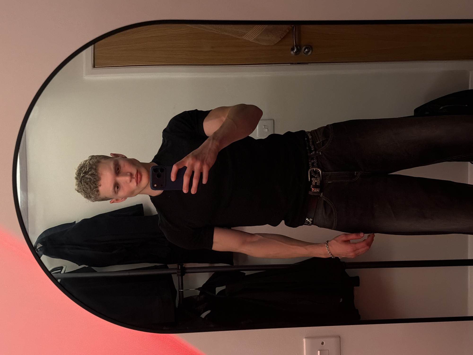 A young, fair-skinned man with short, curly blonde hair and a muscular build is reflected in a large, arched mirror. He is wearing a black t-shirt, dark-wash jeans, and a belt with silver stud detailing. He holds a dark-colored smartphone in his right hand, appearing to take a selfie. The mirror is positioned in what looks like a domestic dressing area, with a clothing rack holding dark garments visible behind him to the left. A white light switch is mounted on the wall to the far left. To the right, a wooden door with a silver handle and a towel hanging on it is reflected. The ambient lighting suggests an indoor setting, possibly a home or a changing room. No specific landmarks are visible that would indicate a location within London, United Kingdom.