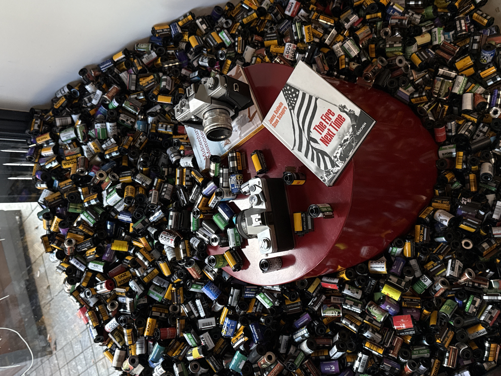 The image presents a collection of film canisters, numbering in the hundreds, primarily brands like Kodak, Fujifilm, and Ilford, scattered on a surface. The canisters appear to be used. Situated atop this pile of film is a vintage Praktica camera, a book titled "The Fire Next Time" by James Baldwin with photography by Steve Schapiro, and a burgundy record-shaped stand. Based on the contents of the image, there is no information to place the image in Belgrade, Serbia.
