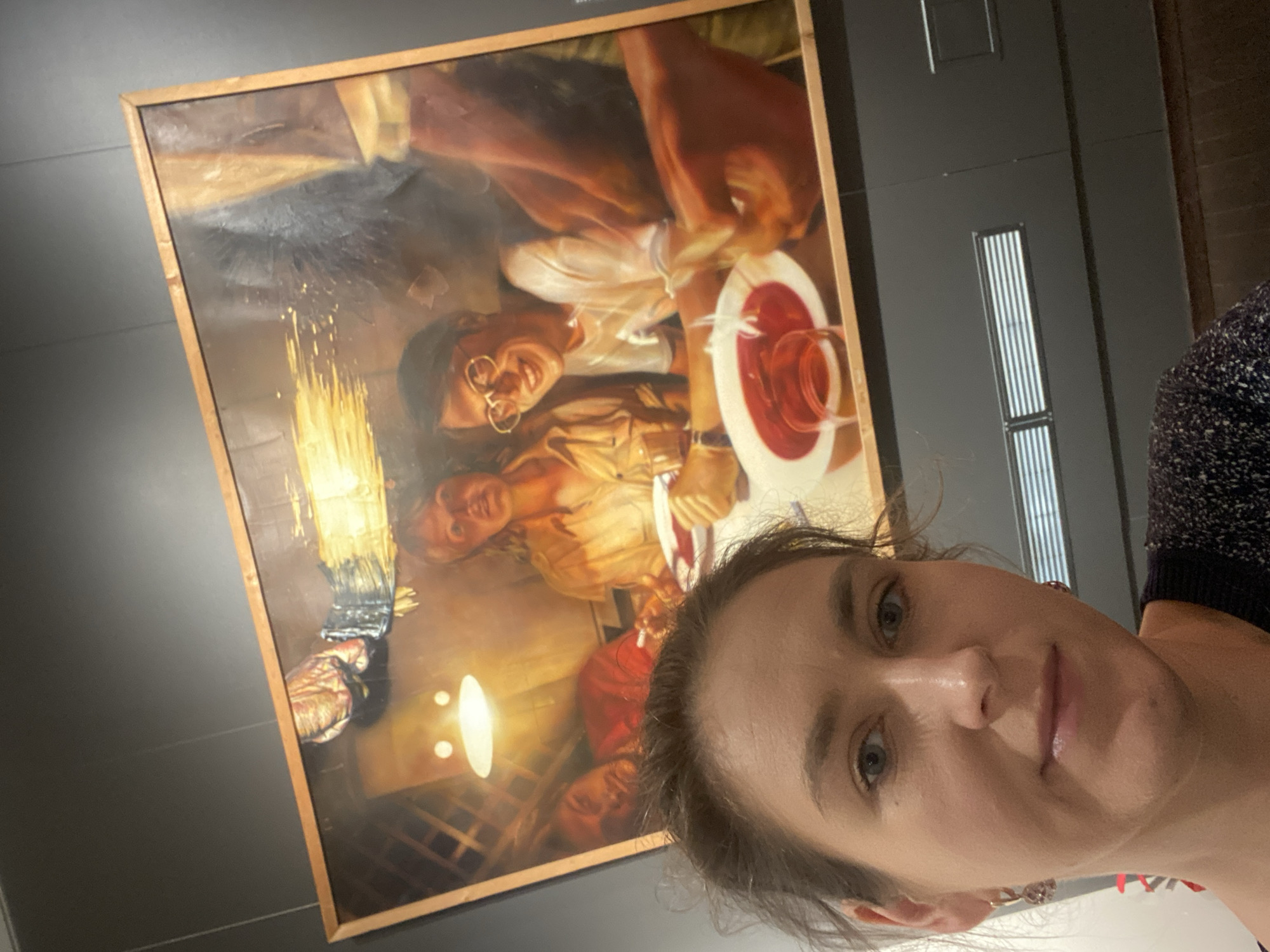 The image shows a woman taking a selfie in front of a large oil painting. The painting depicts a group of people gathered around a table. The woman's face is partially visible in the foreground, with her eyes looking directly at the camera. The painting is framed and hangs on a wall in what appears to be an art gallery or museum. The background of the painting shows a dimly lit interior scene with several figures. One figure in the foreground of the painting is a man wearing glasses and smiling. The lighting in the scene is warm and artificial. The overall impression is that the woman is visiting an art exhibition in Yekaterinburg, Russia.