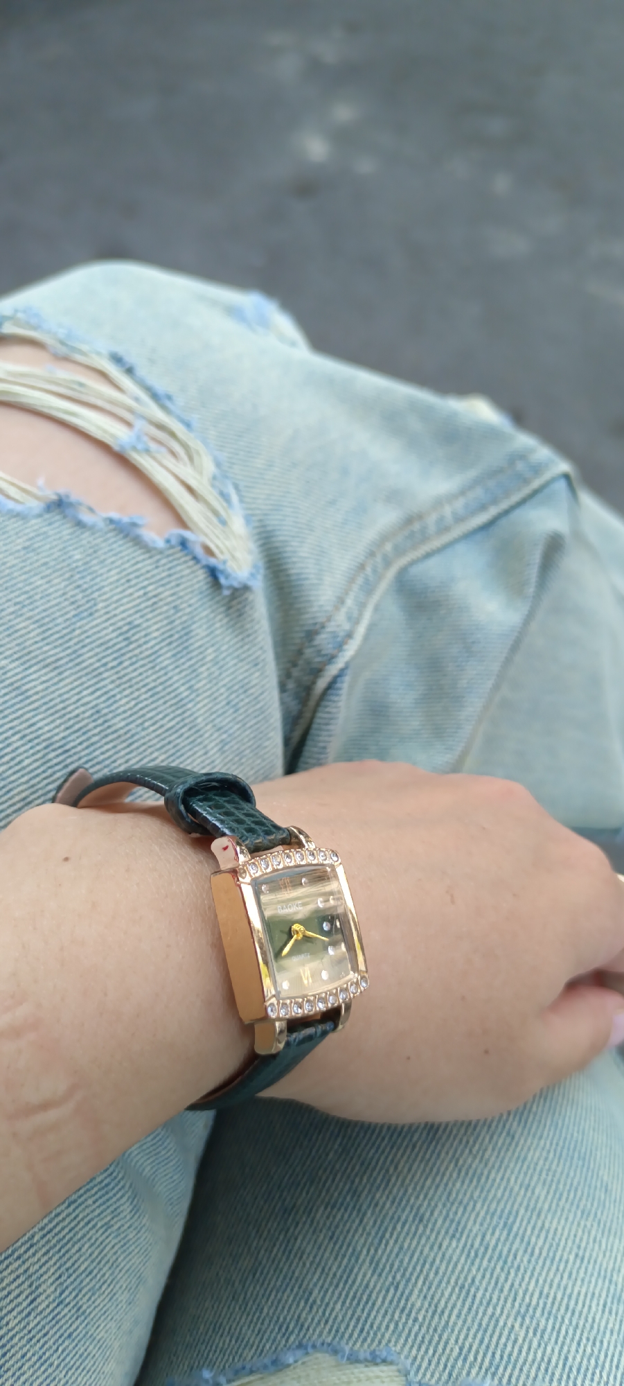 The image shows a close-up of a person's wrist wearing a gold-colored square watch with a dark green band. The watch face is green and has Roman numerals. The bezel of the watch is adorned with small clear stones. The person is wearing ripped light-wash denim jeans. The background is blurred and appears to be an outdoor setting with a greyish-black surface, possibly pavement. The image does not contain any information to suggest it is located in Fergana City, Uzbekistan.